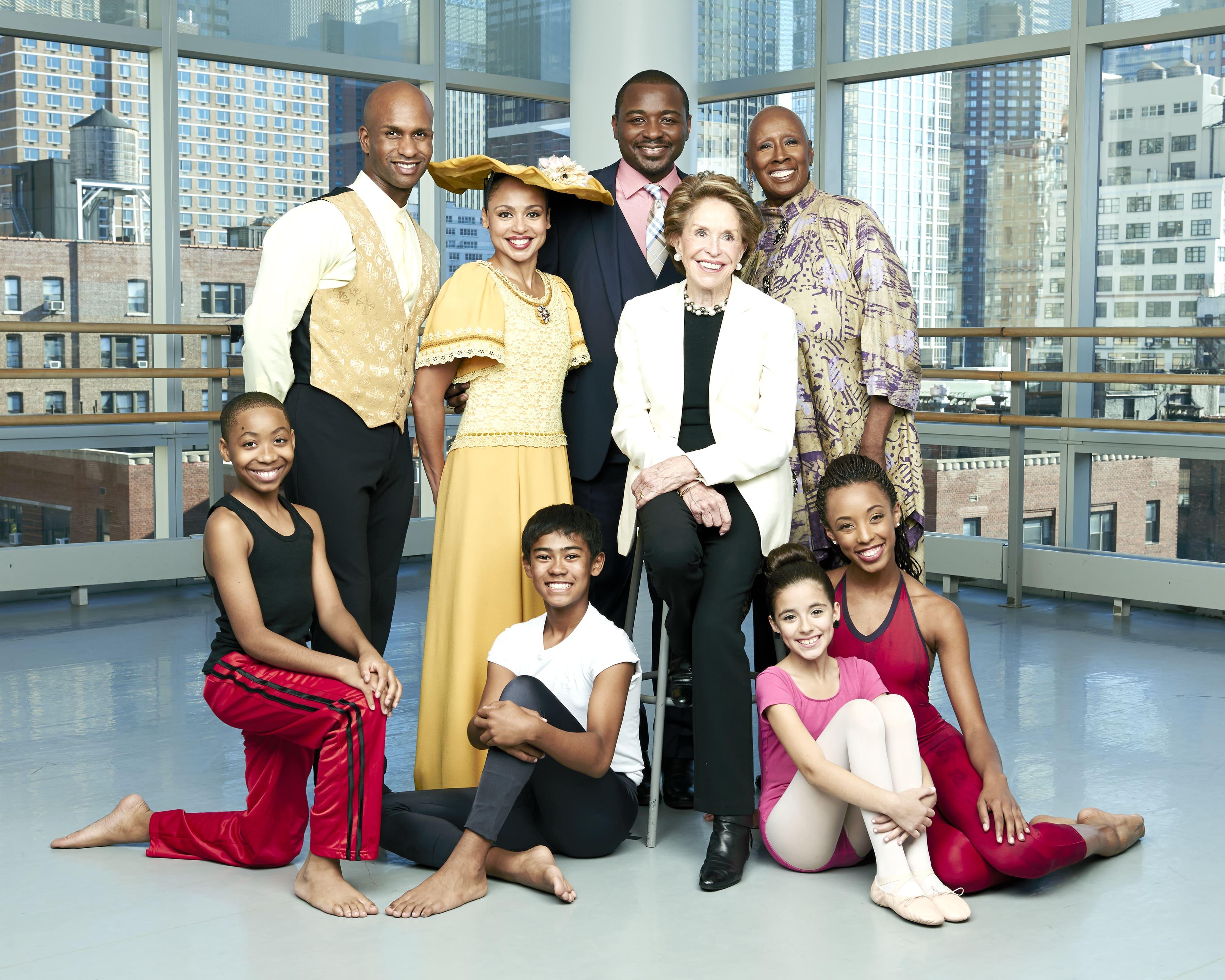 Group photo featuring Joan Weill, Robert Battle, Judith Jamison, Glenn Allen Sims, Linda Celeste Sims, and students from The Ailey School. They are posed in a bright, spacious studio with large windows showing a cityscape. Joan Weill is seated in the center, surrounded by the others who are standing and kneeling, all smiling. Photo by Andrew Eccles, 2014.