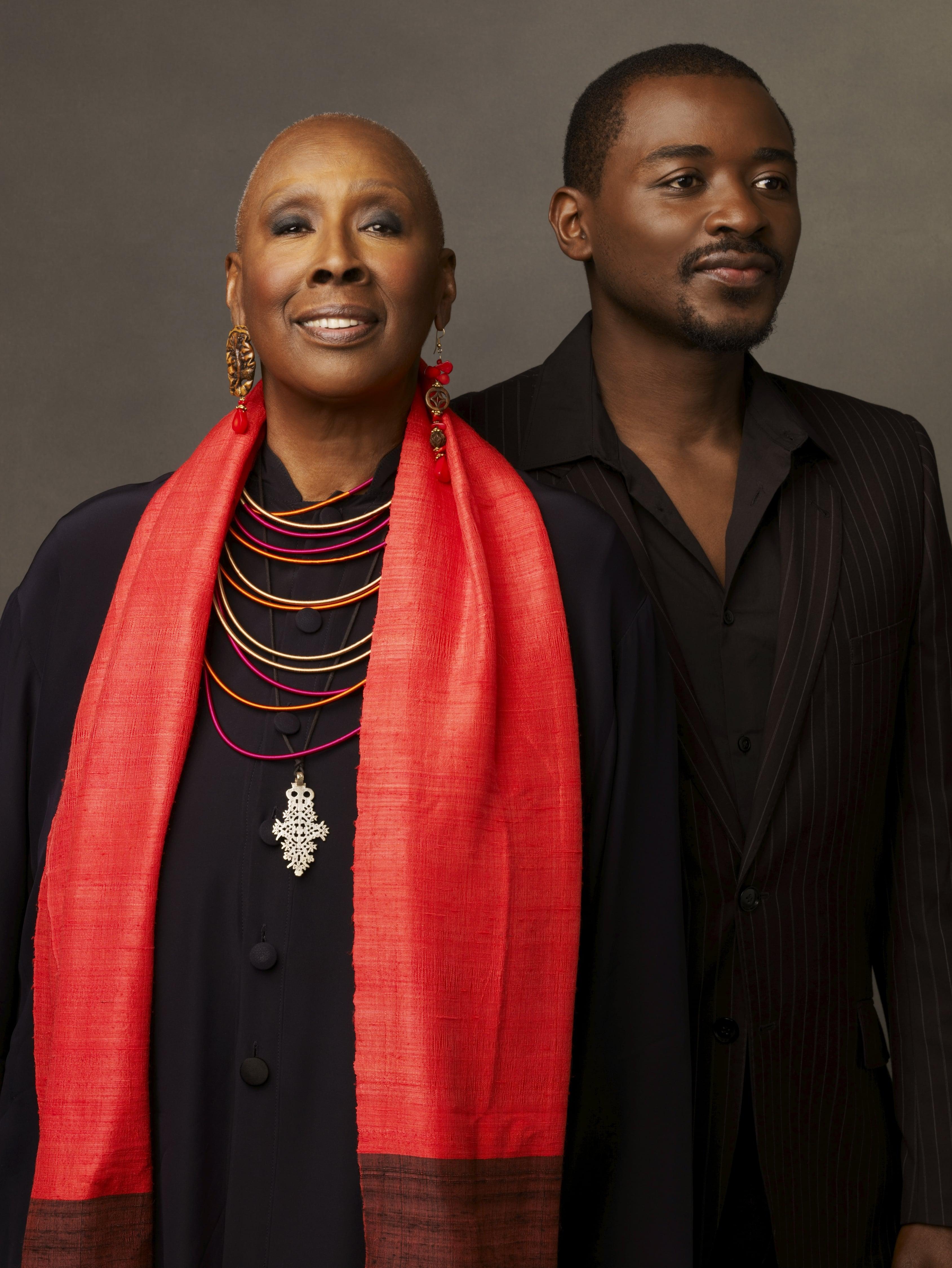 Judith Jamison and Robert Battle standing side by side, both smiling. Jamison is wearing a dark outfit with a red scarf and layered necklaces, while Battle is in a dark pinstriped suit. The background is a plain, neutral color. Photo by Andrew Eccles, 2011.