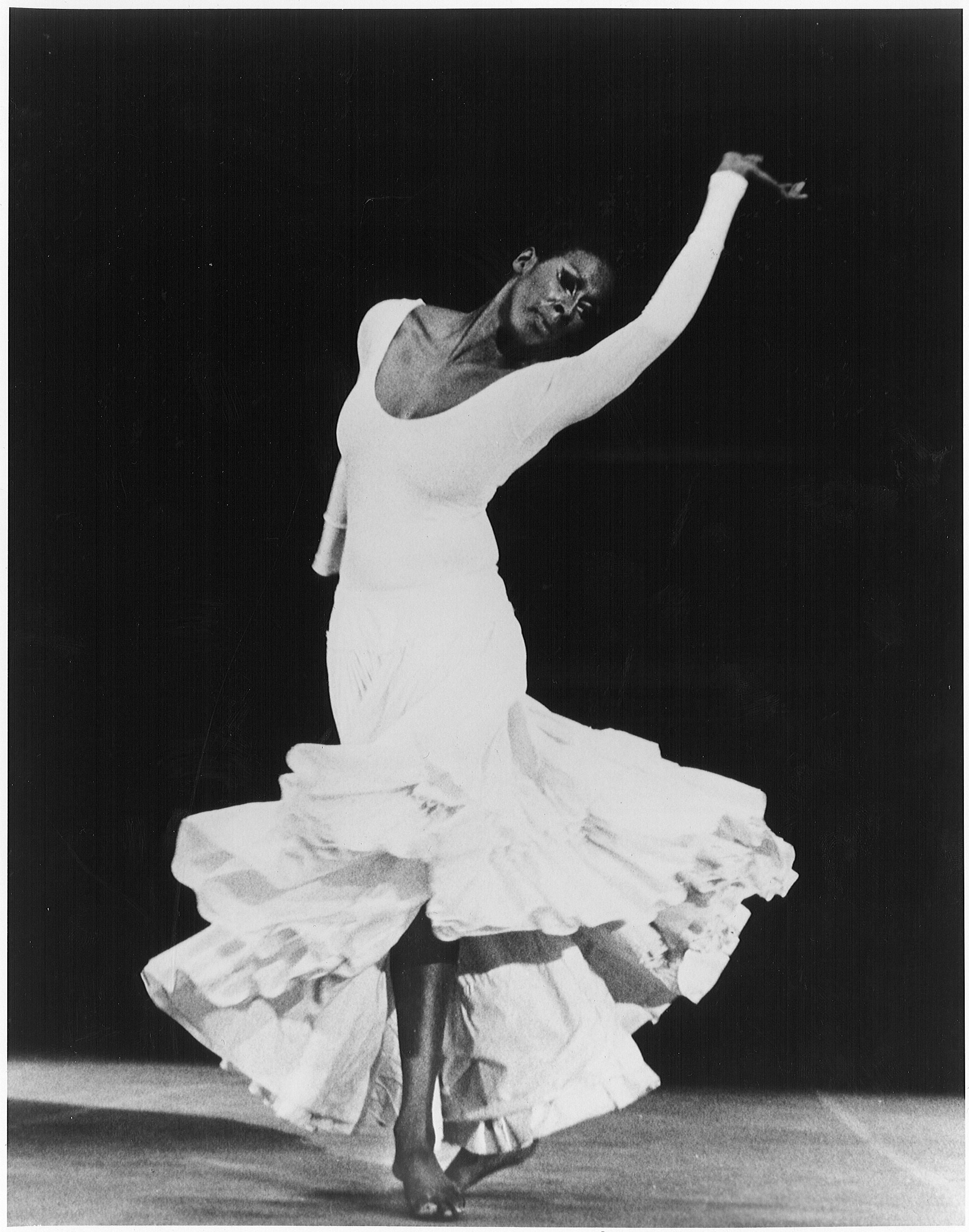 Judith Jamison performing in Alvin Ailey's "Cry," wearing a long white dress with a full skirt. She is captured mid-movement with one arm raised and her head tilted back, exuding emotion and grace. The background is dark, emphasizing her dynamic pose. Photo by R. Faligant.