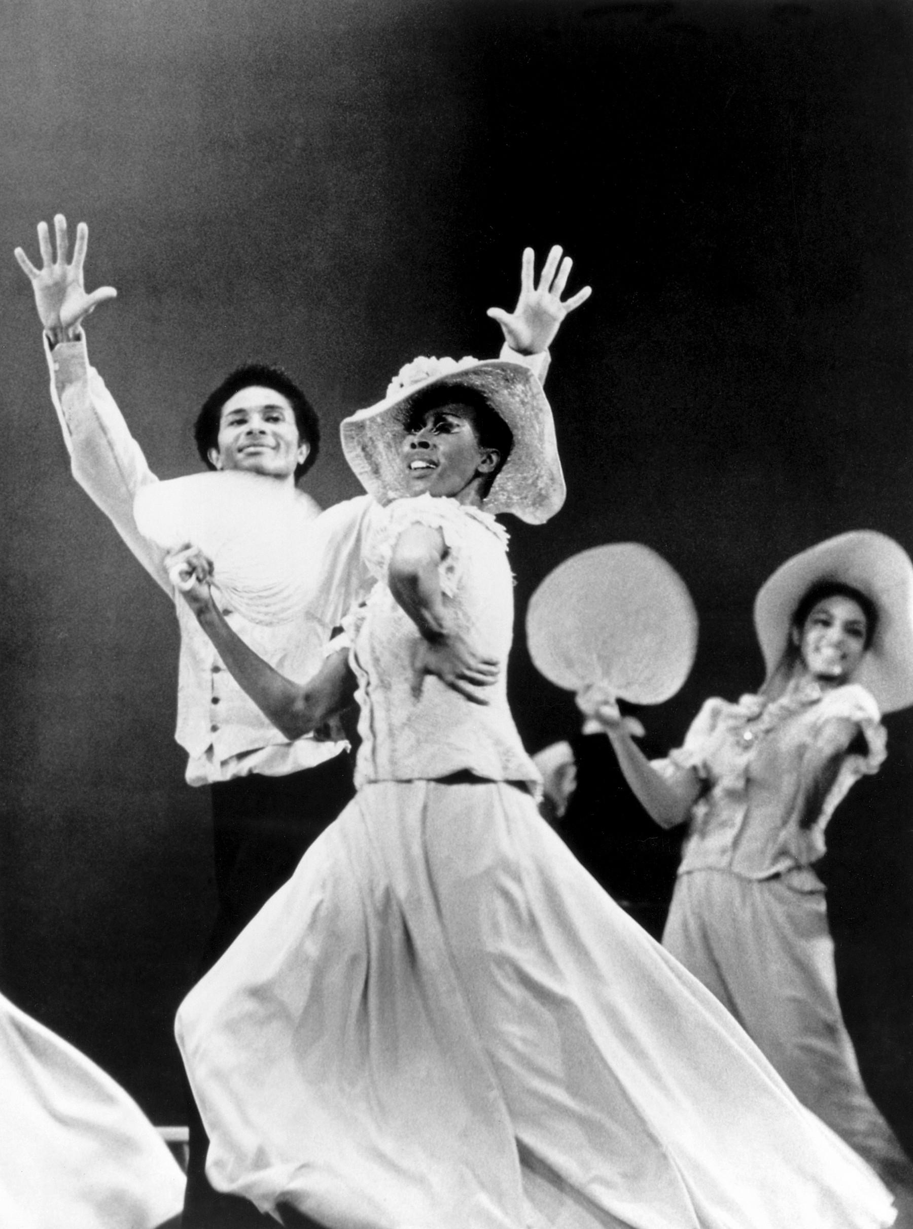 Judith Jamison performing in Alvin Ailey's "Revelations" in 1965, captured by George Kalinsky. She is wearing a light-colored dress and a hat, holding a fan in one hand. Her expression and pose exude energy and passion, with her arm raised high. Other dancers in similar attire are visible in the background, contributing to the dynamic and lively atmosphere of the performance.