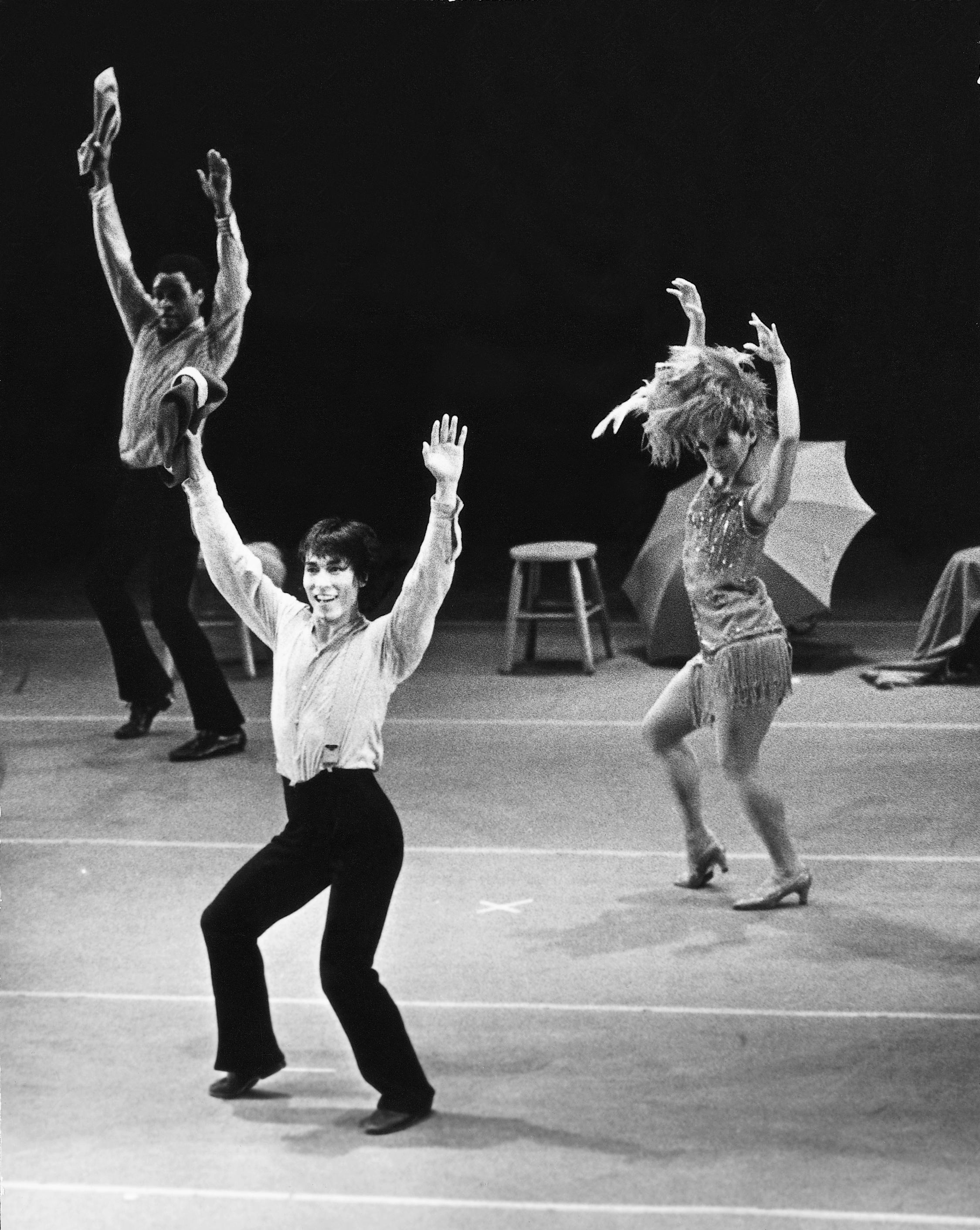 This photograph, taken in 1972, features Masazumi Chaya, Melvin Jones, and Mari Kajiwara performing in Alvin Ailey's "Blues Suite." Courtesy of the Ailey Archives, the image captures a lively moment during the dance. Chaya, in the foreground, raises his arms with a joyful expression, while Jones and Kajiwara, in the background, also display exuberant poses. Jones holds his hat high, and Kajiwara, wearing a fringed outfit, adds a sense of movement and energy to the scene. The stage is set with simple props, 