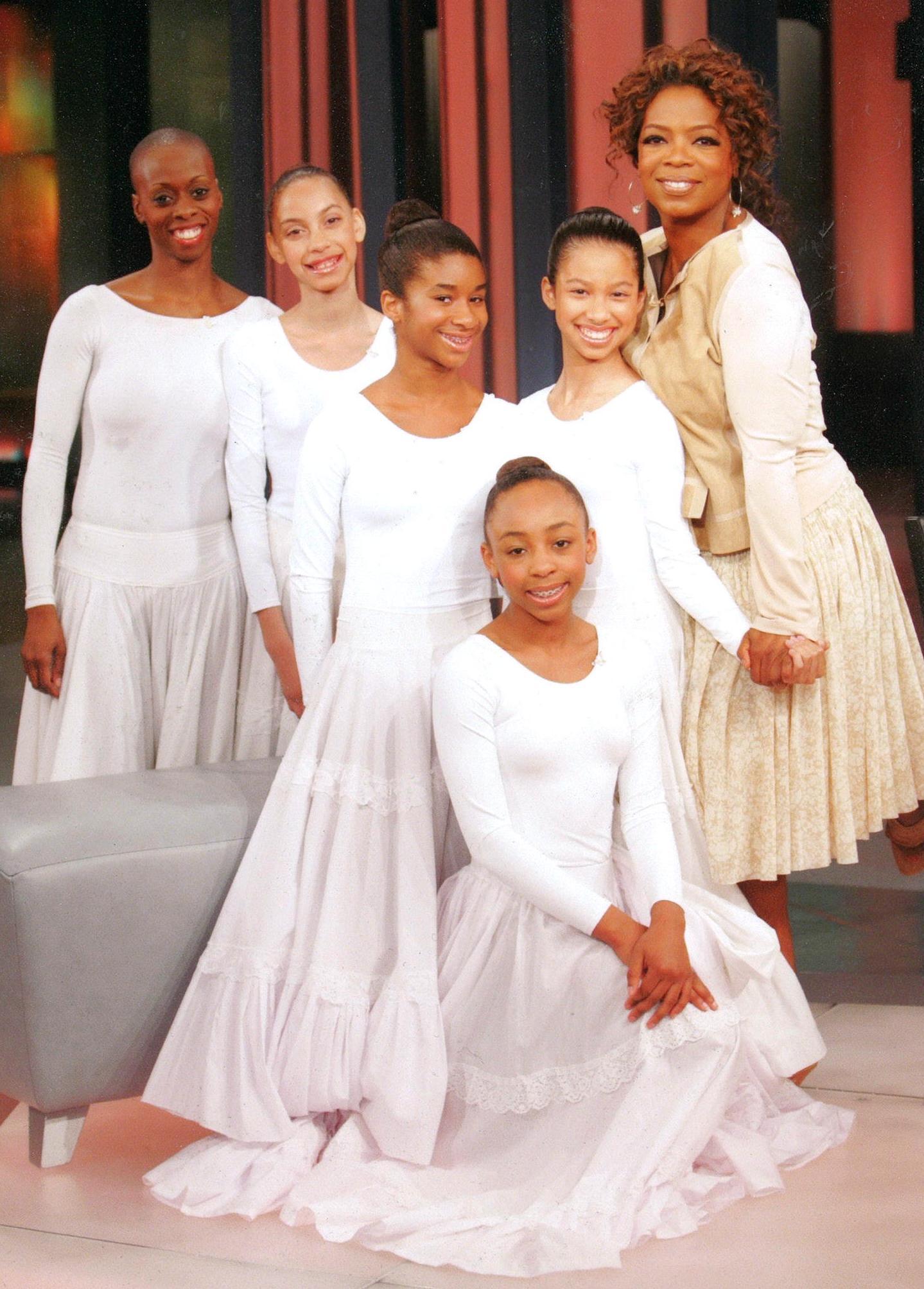  This image features students from The Ailey School along with D. Smallwood from the Alvin Ailey American Dance Theater, posing with Oprah Winfrey during a taping of "The Oprah Winfrey Show" in 2007. The students and dancer are dressed in white, reminiscent of the costumes often seen in performances of "Revelations," one of Alvin Ailey's most iconic works. Oprah Winfrey, a well-known supporter of the arts, is seen smiling and holding hands with one of the students, highlighting the connection and support be