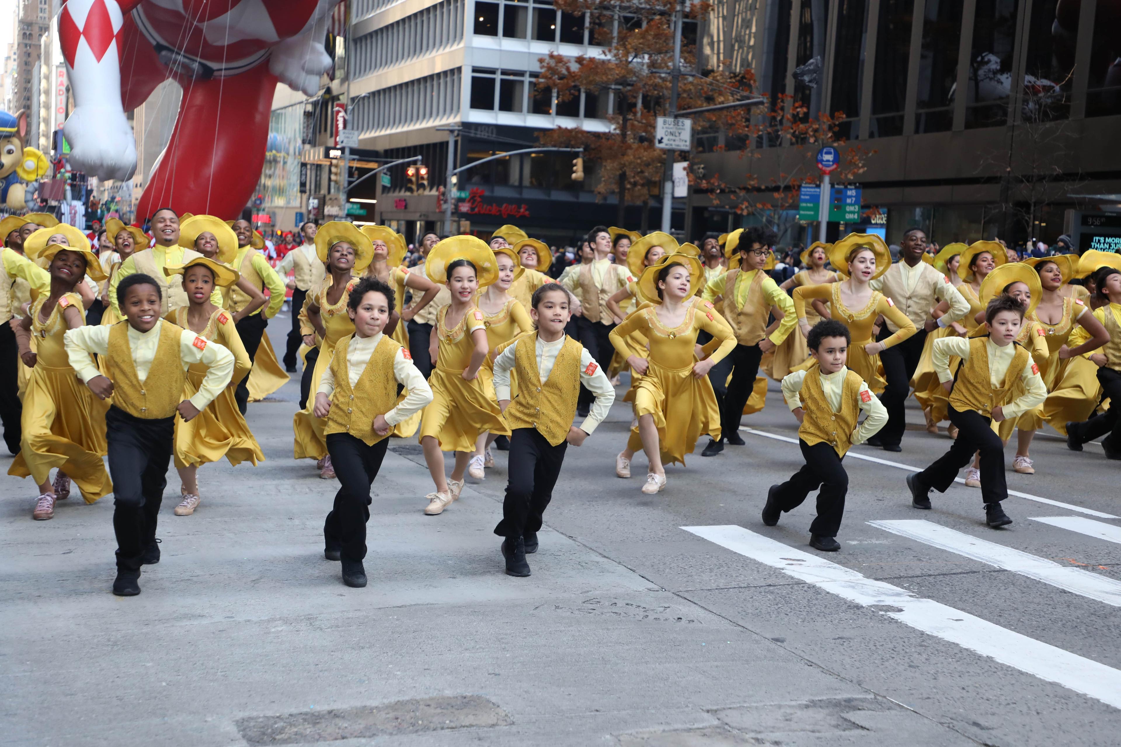 This image captures a vibrant and energetic moment from the Macy's Thanksgiving Day Parade in 2019. The students from The Ailey School are seen dancing in coordinated golden costumes, bringing joy and enthusiasm to the streets. Their performance is full of spirit, showcasing the talent and dedication nurtured at The Ailey School. The photograph, taken by Mariah Gravelin, highlights the school's involvement in this iconic parade, reflecting the school's commitment to community engagement and the celebration 