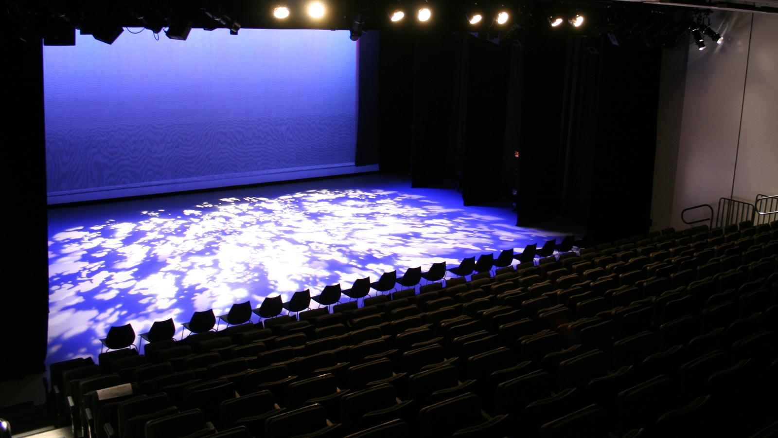 An empty theater. The stage is lit in dark blue and white. The chairs are dark.