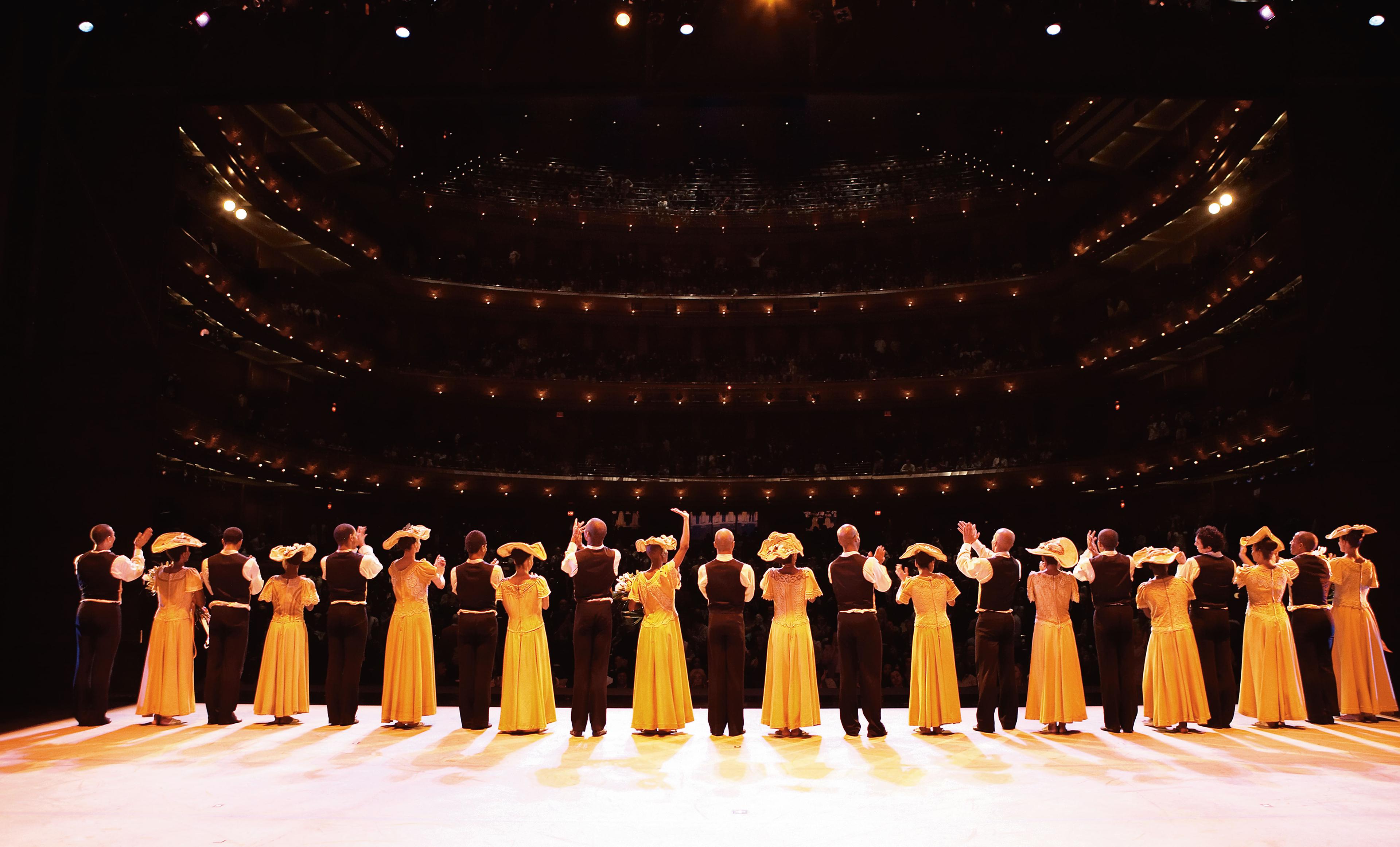 A line of dancers stands on stage with their hands held, ready to bow. The dancers alternate men and women. The women are in yellow dresses with hats. The men are in black dress pants, vests, white shirts.