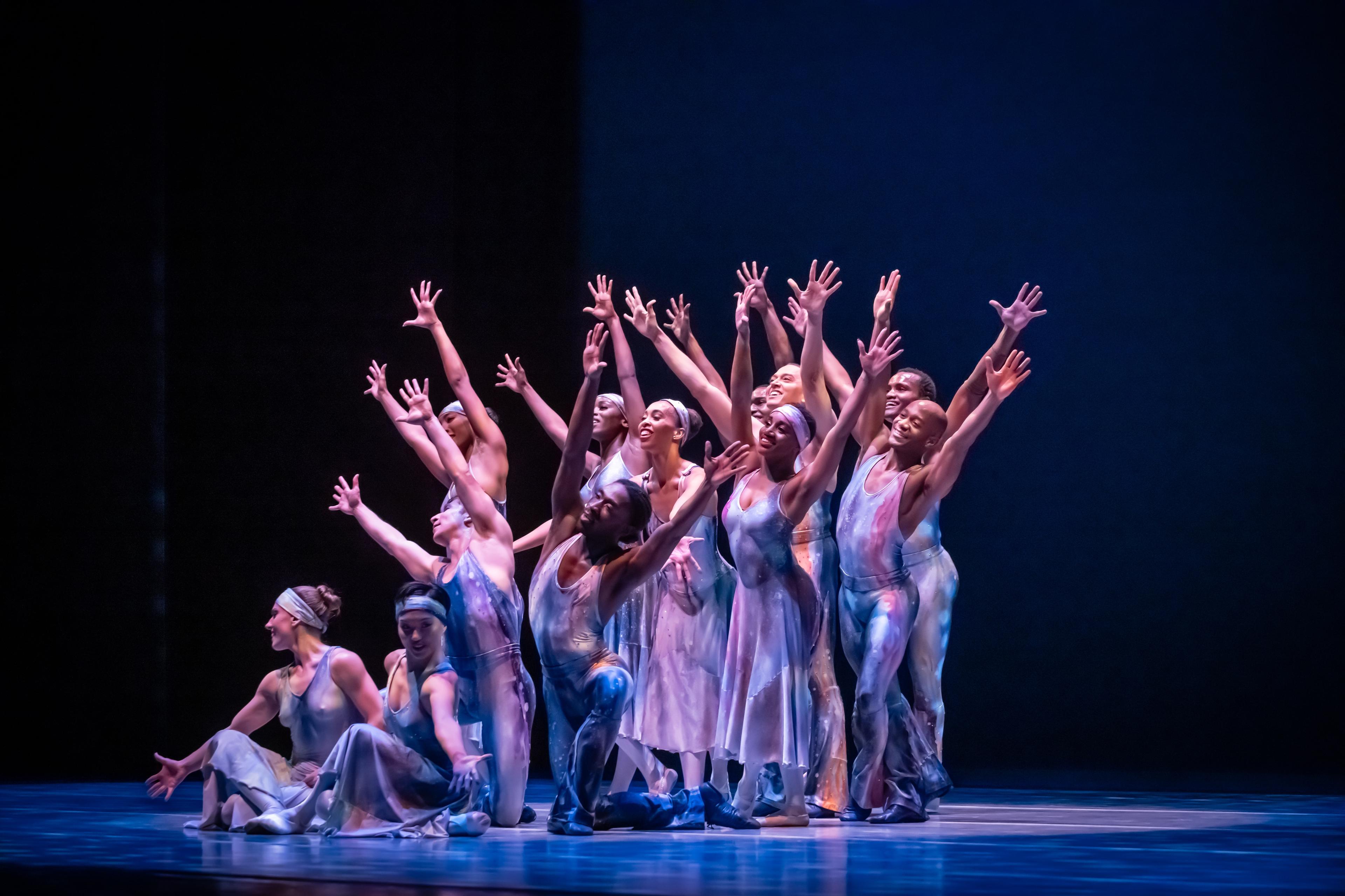 A group of dancers on stage in blue/purple dresses. They are huddled with their arms outstretched