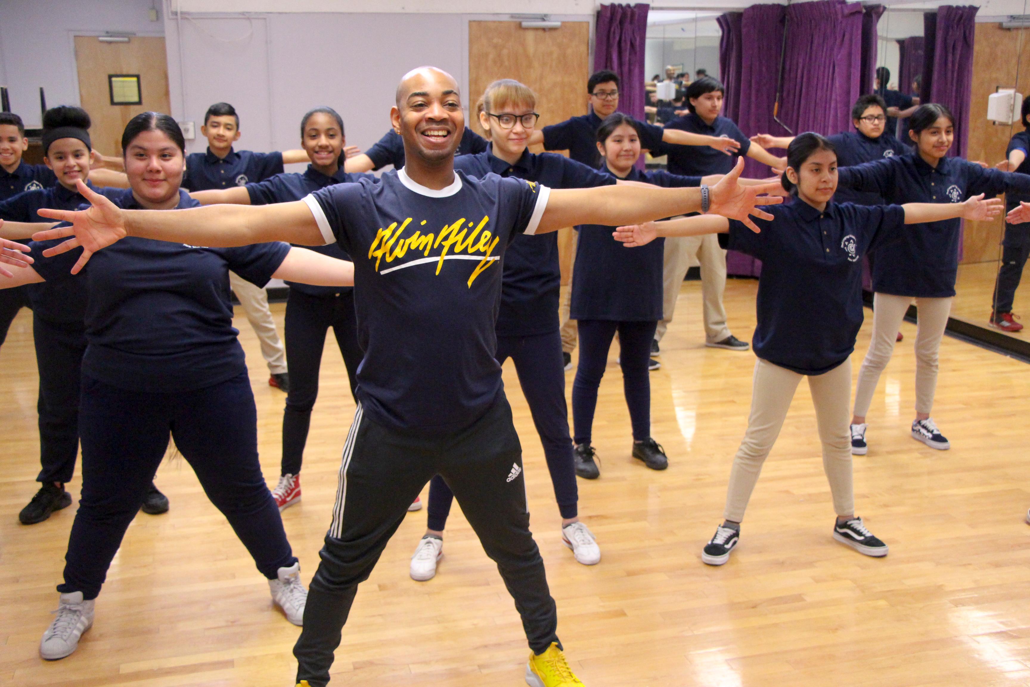 A teacher with students in a dance studio. They are all wearing navy shirts and have their arms outstretched.