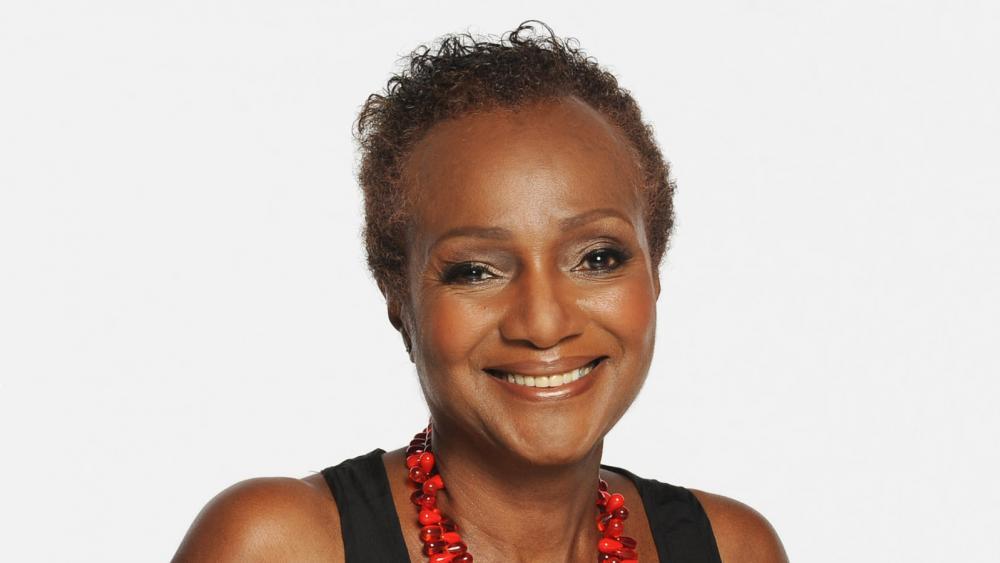 Portrait of Sylvia Waters wearing a black top and a red beaded necklace. She is smiling and looking directly at the camera. The background is plain and white.