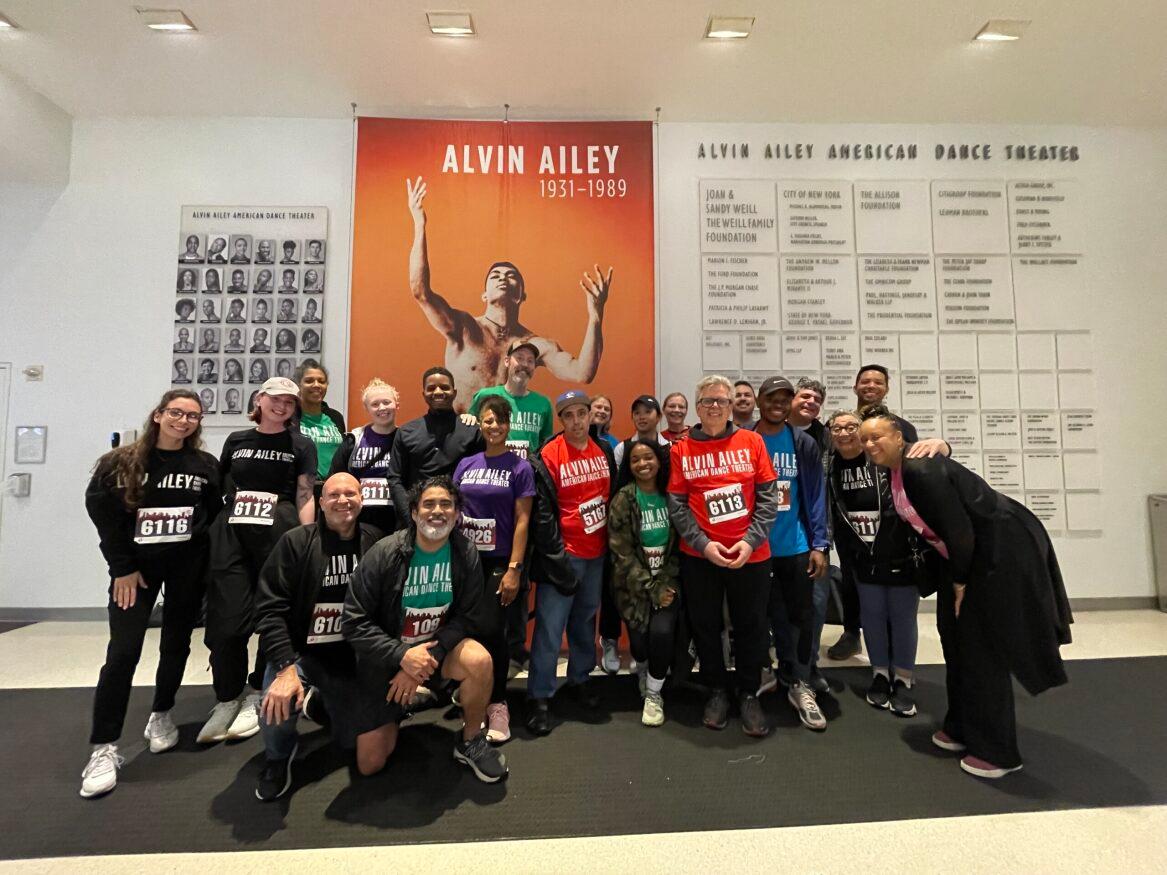 A team stands in front of an Alvin Ailey poster. They are all wearing race shirts and bibs