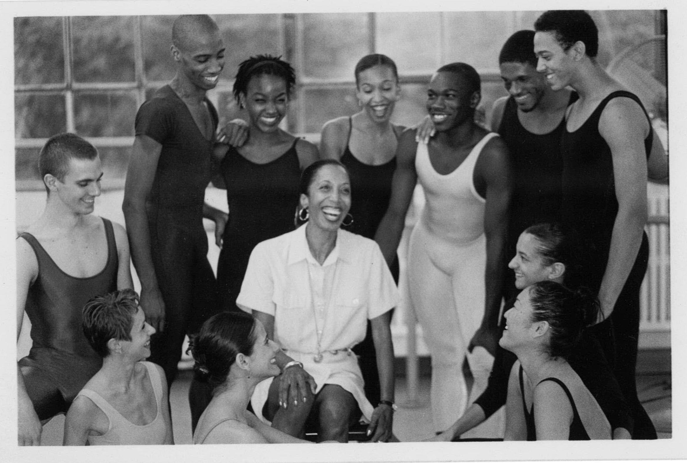 A group of dancers smiling together in a studio. The photo is black and white