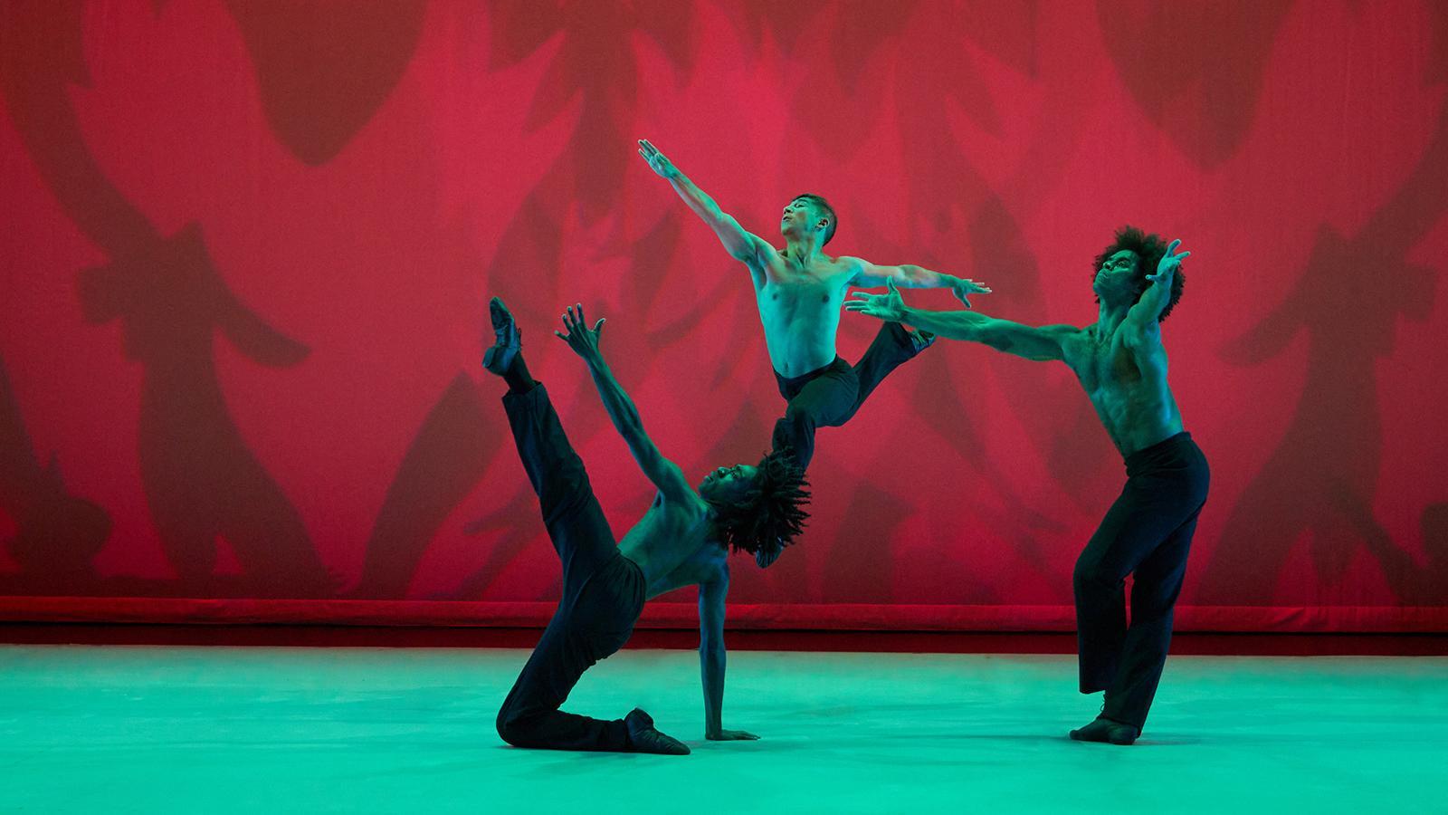 Three male dancers perform on stage against a red backdrop with abstract shadows. The dancers are shirtless, wearing black pants, and are posed dynamically with one dancer on the floor, one mid-air, and another extending an arm. The stage is lit with green and red lighting.