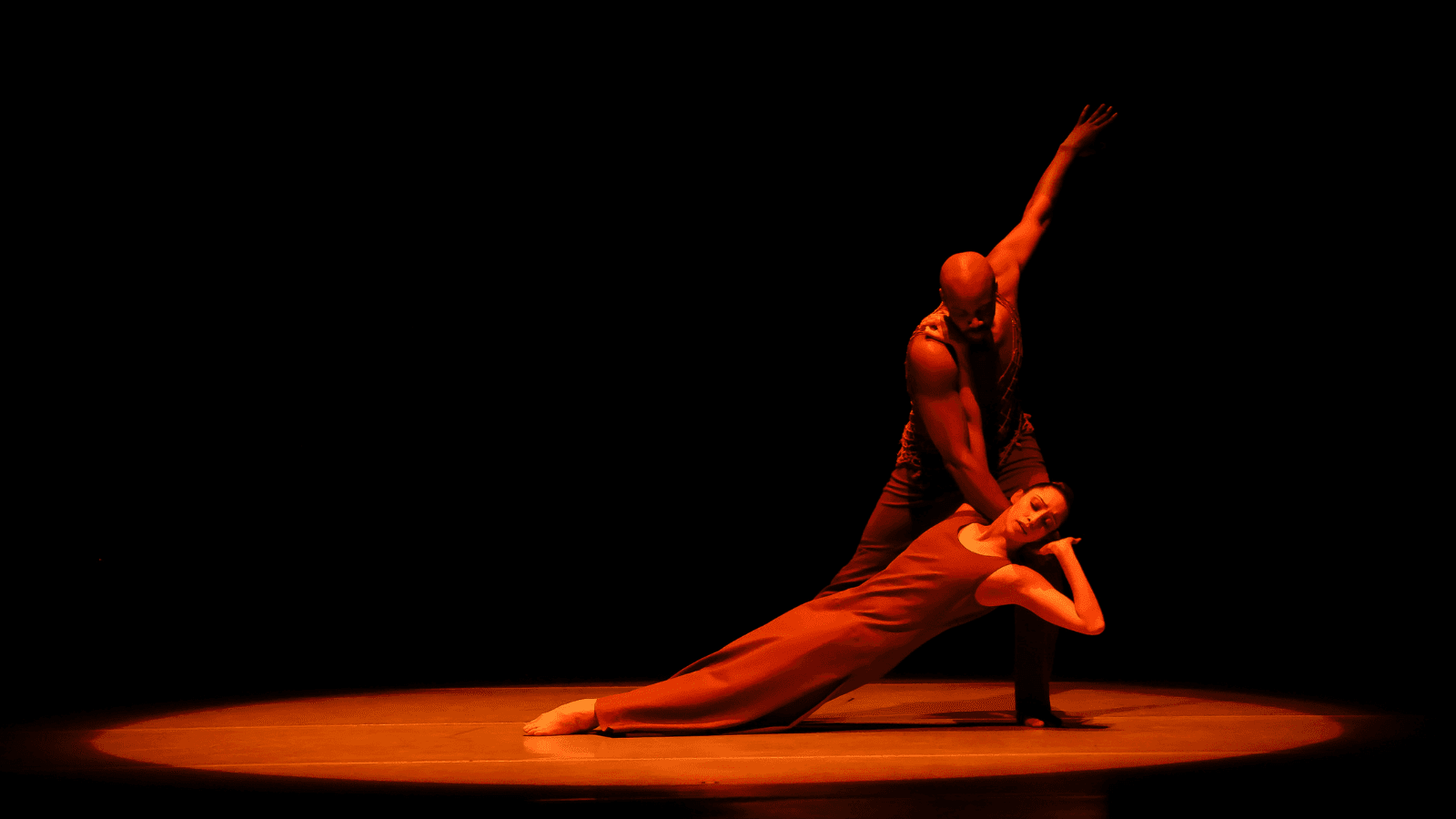 Two dancers perform on stage under a spotlight. One dancer is standing with an arm extended upward, while the other is leaning back gracefully, supported by the standing dancer. Both are dressed in red costumes and the stage is dark except for the warm spotlight on the performers.