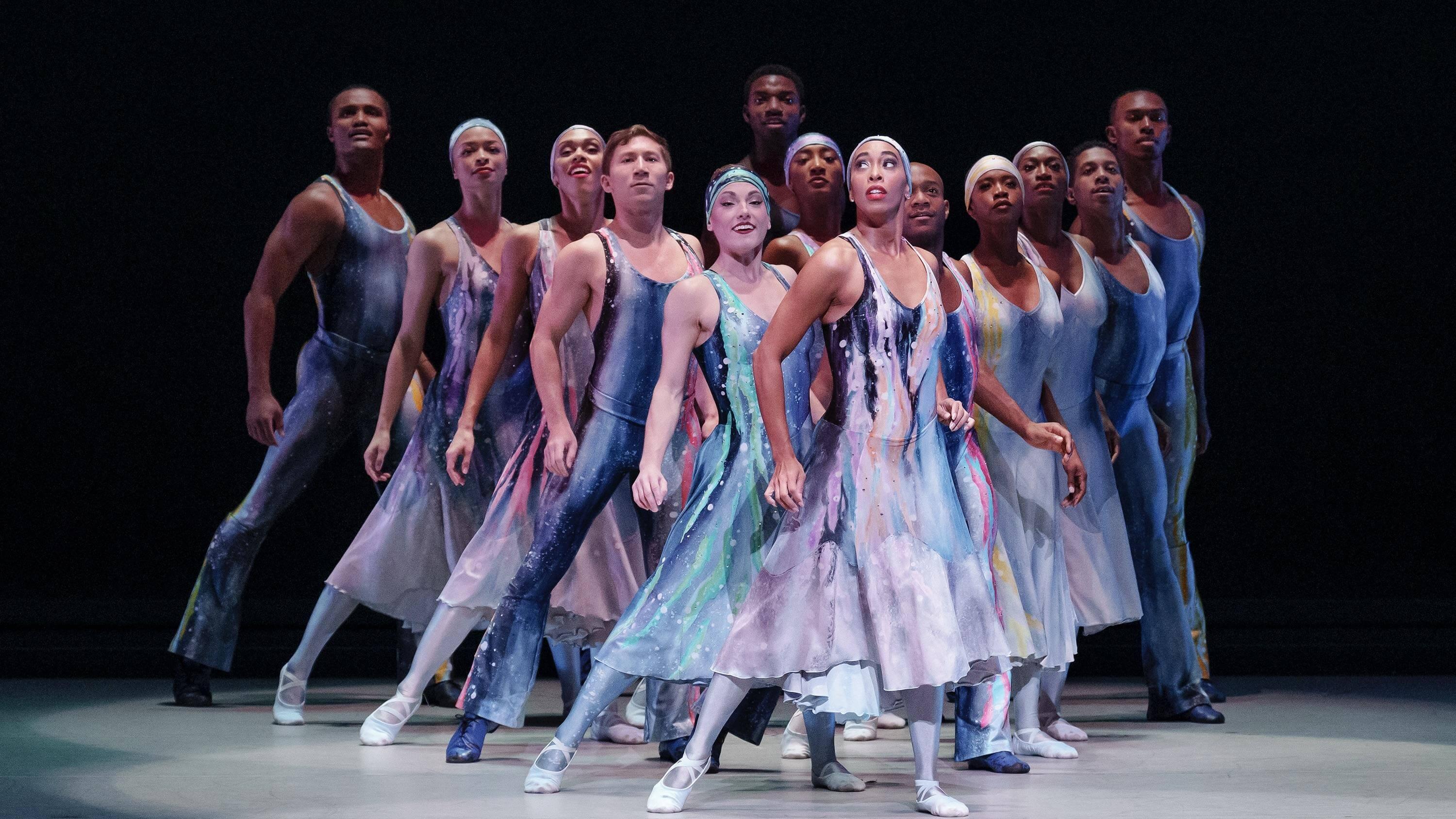 A group of dancers on stage in blue gradient costumers stand in a V shape all looking up.