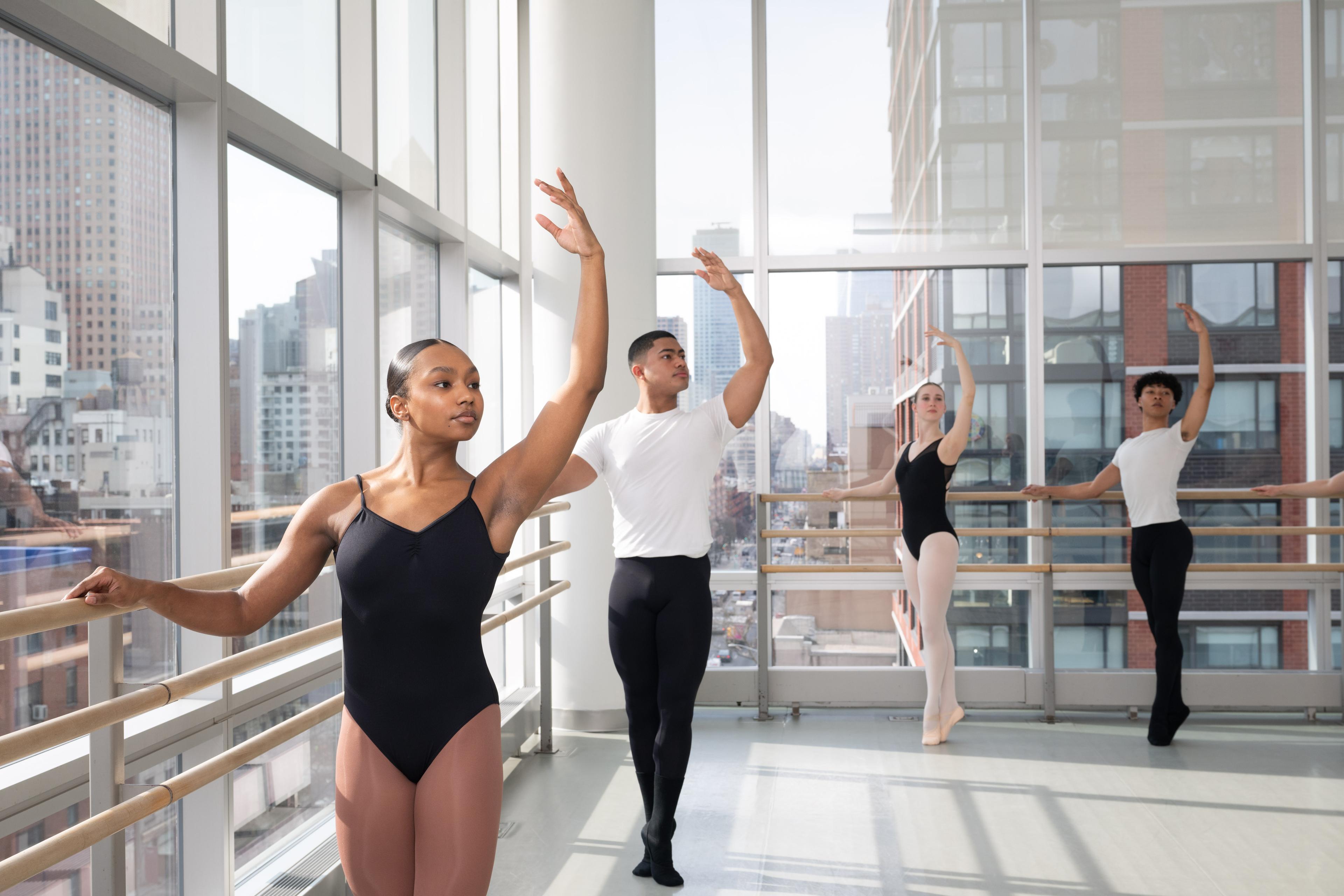 A group of students at a ballet bar in a dance studio. They have one hand on the bar and the other above their heads. The girls are in black leotards and the boys in black pants and white tee shirts.