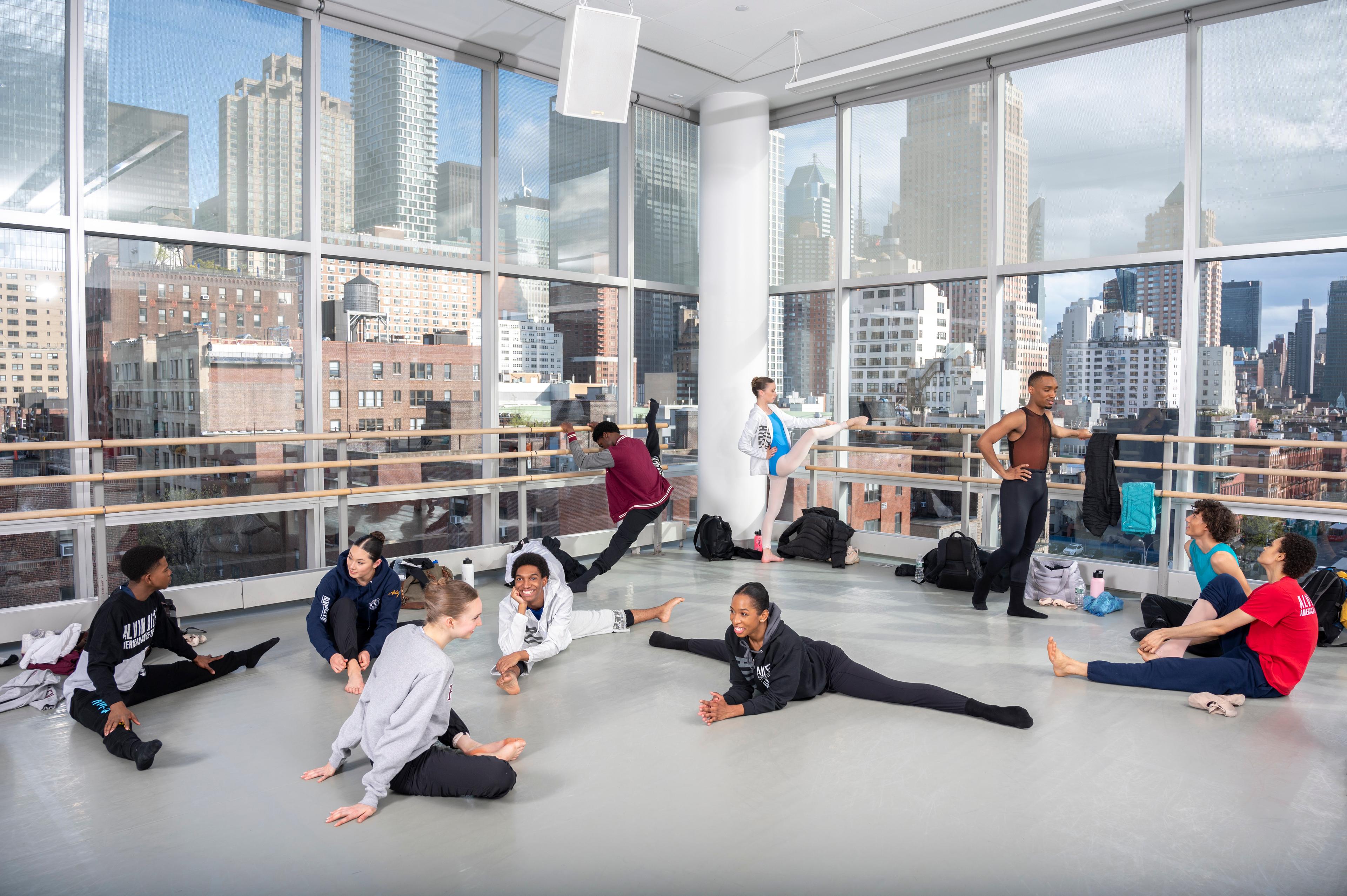 a group of students at The Ailey School hanging out and stretching in a dance studio with floor to ceiling windows.