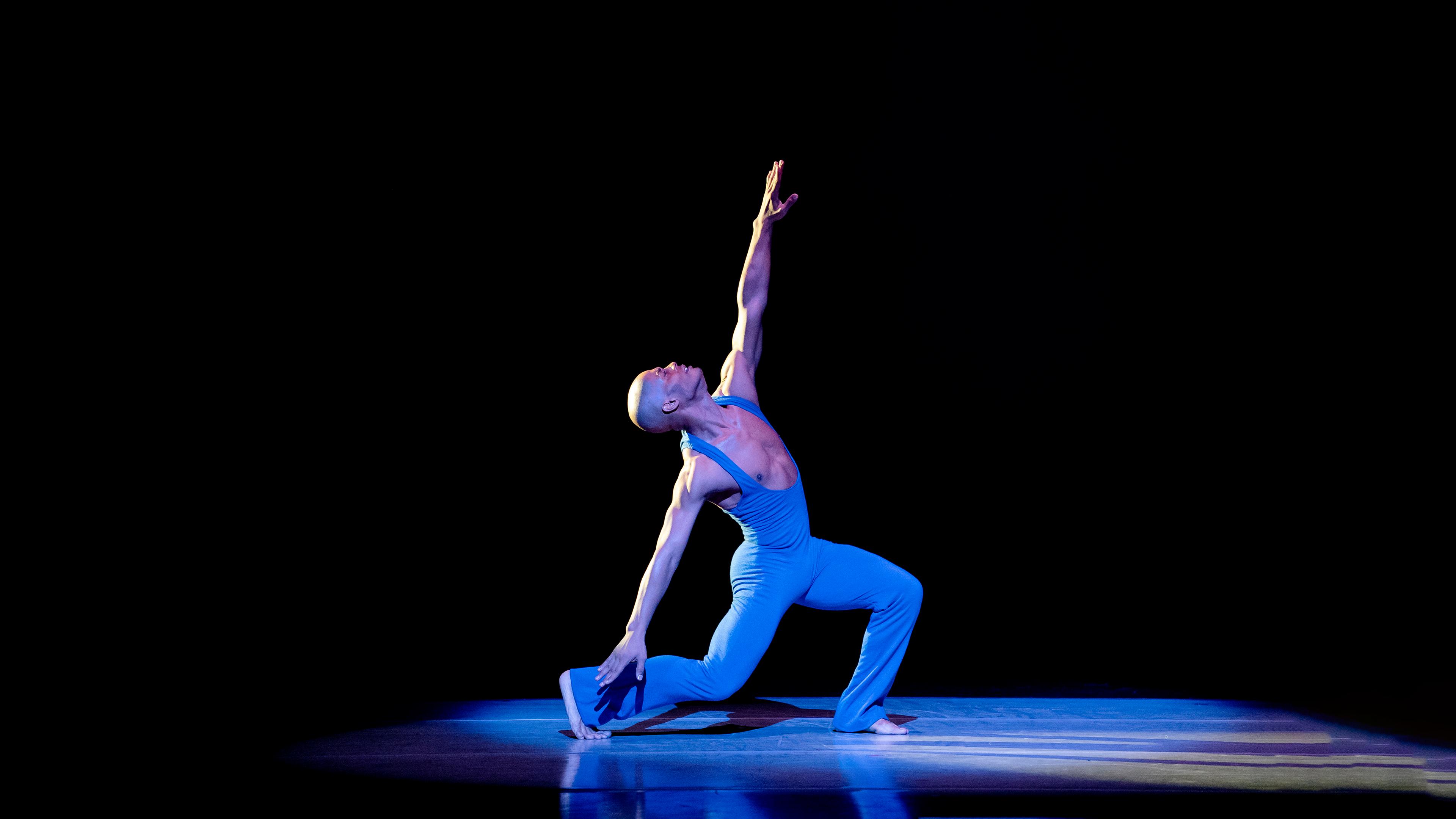 A dancer wearing a blue leotard dancing. The background is black.