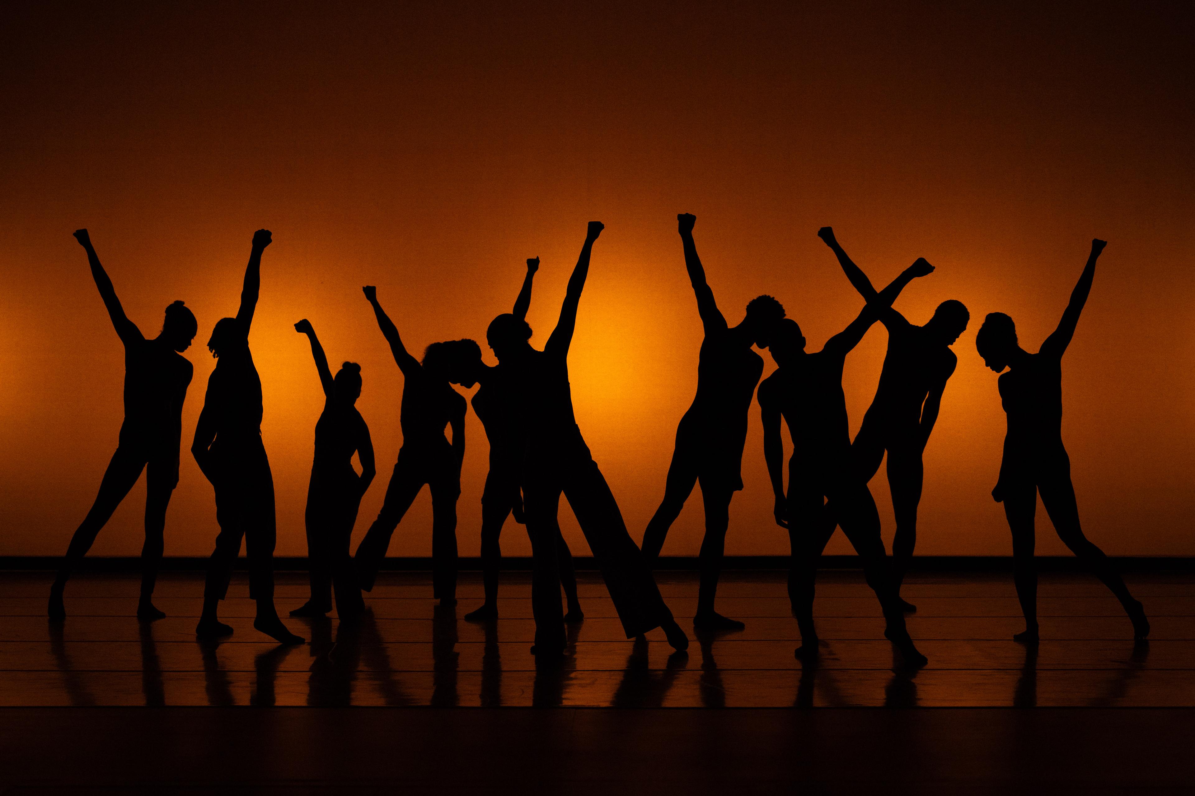 A silhouetted group of dancers stand on stange with one arm raised into the air.