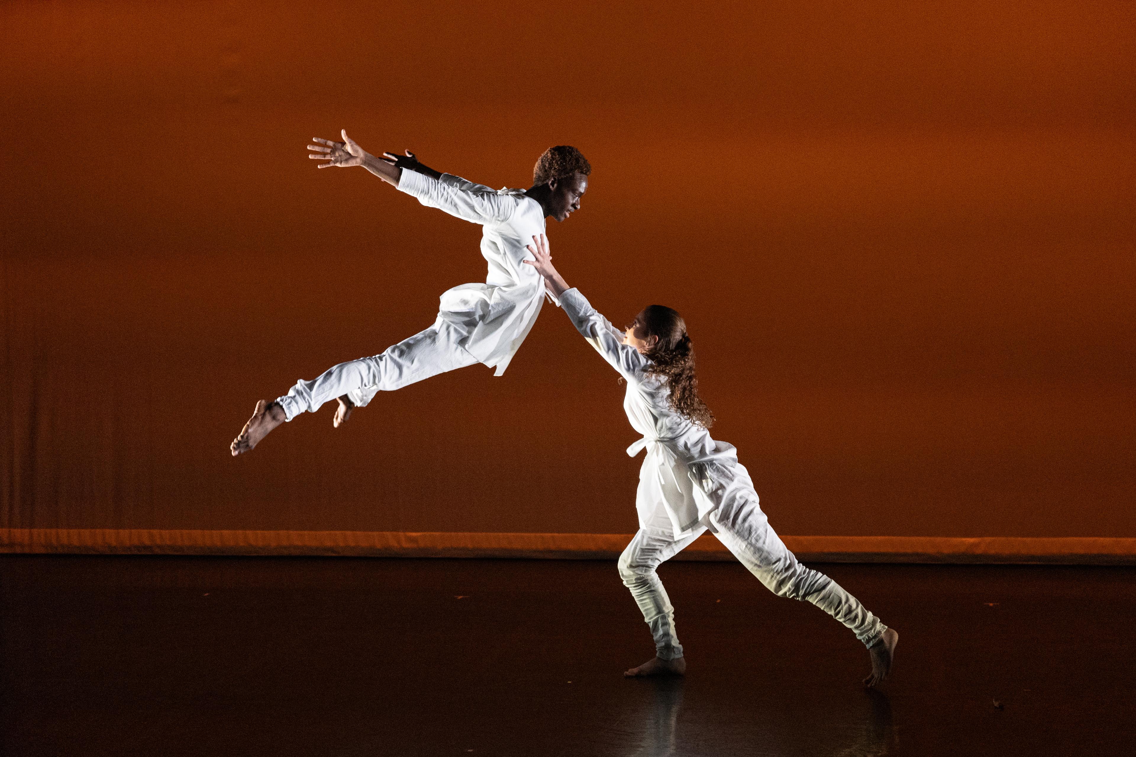 Two dancers dressed in white, on jumping up in the air towards the second one standing nearby