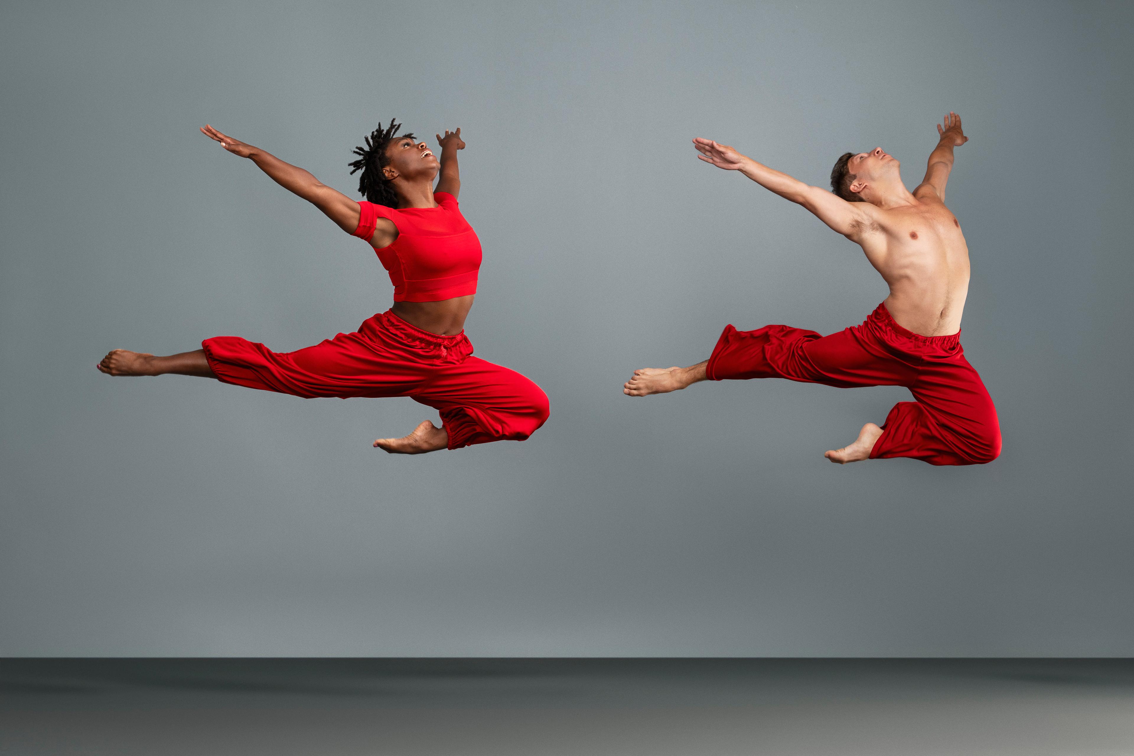 There are two dancers jumping in the air. Both are wearing red. They are in a studio with grey walls.