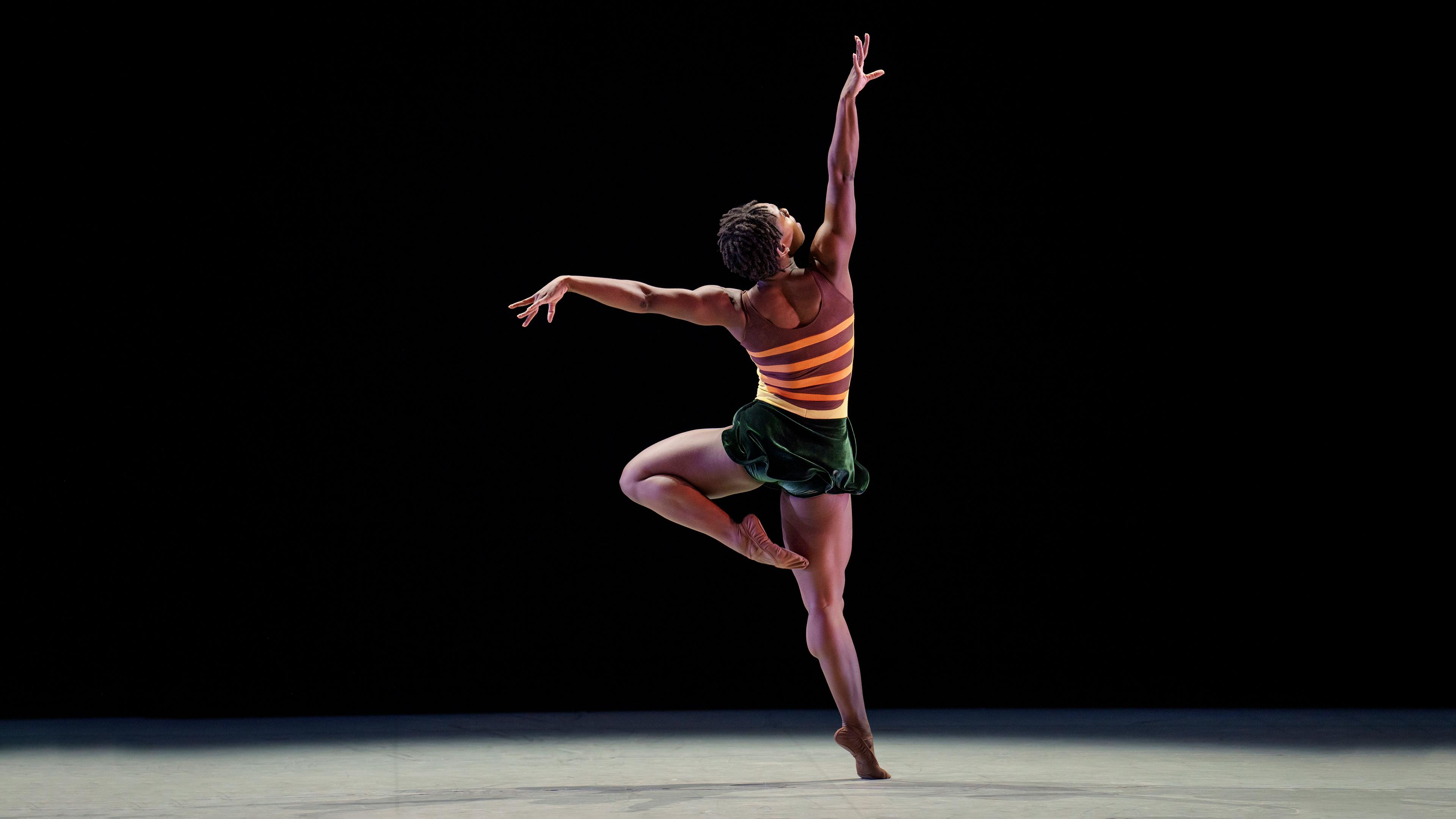 A woman with her back to the audience stands on one leg reaching one had up to the sky.