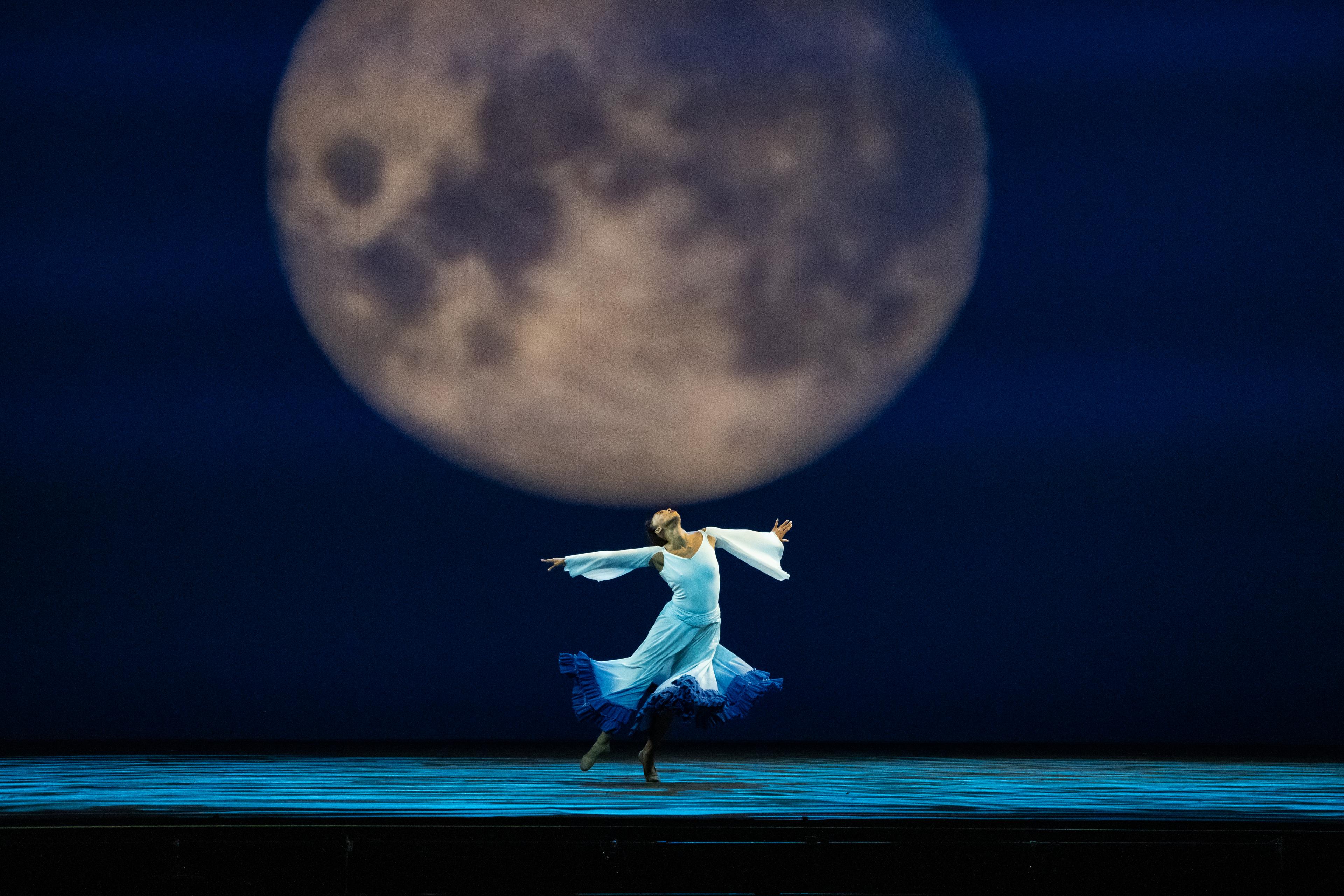 A woman in a white dress spins around in front of a backdrop of a full moon.