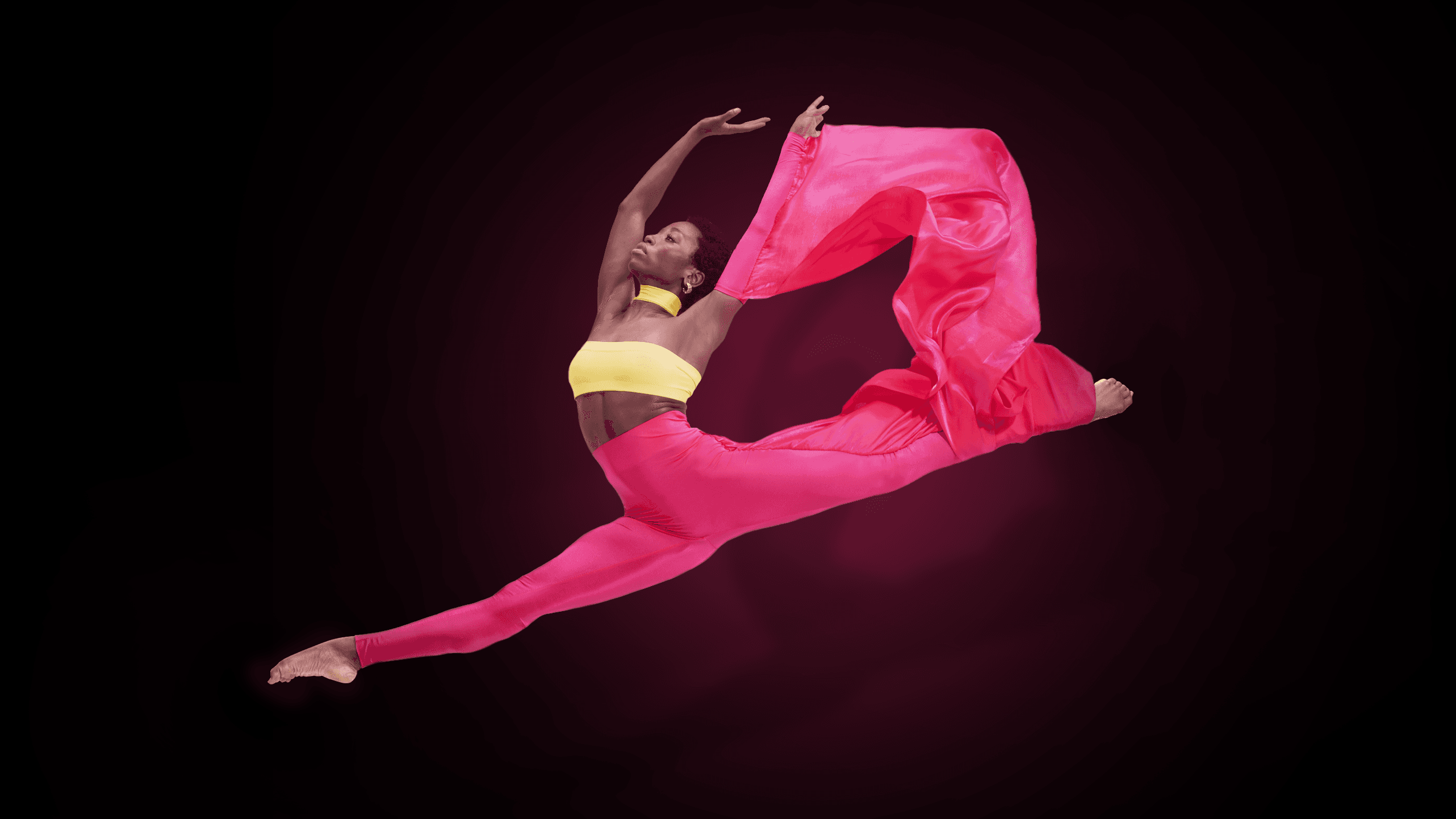 Dancer against a black background. They are leaping in the air and wearing a bright pink ensemble.