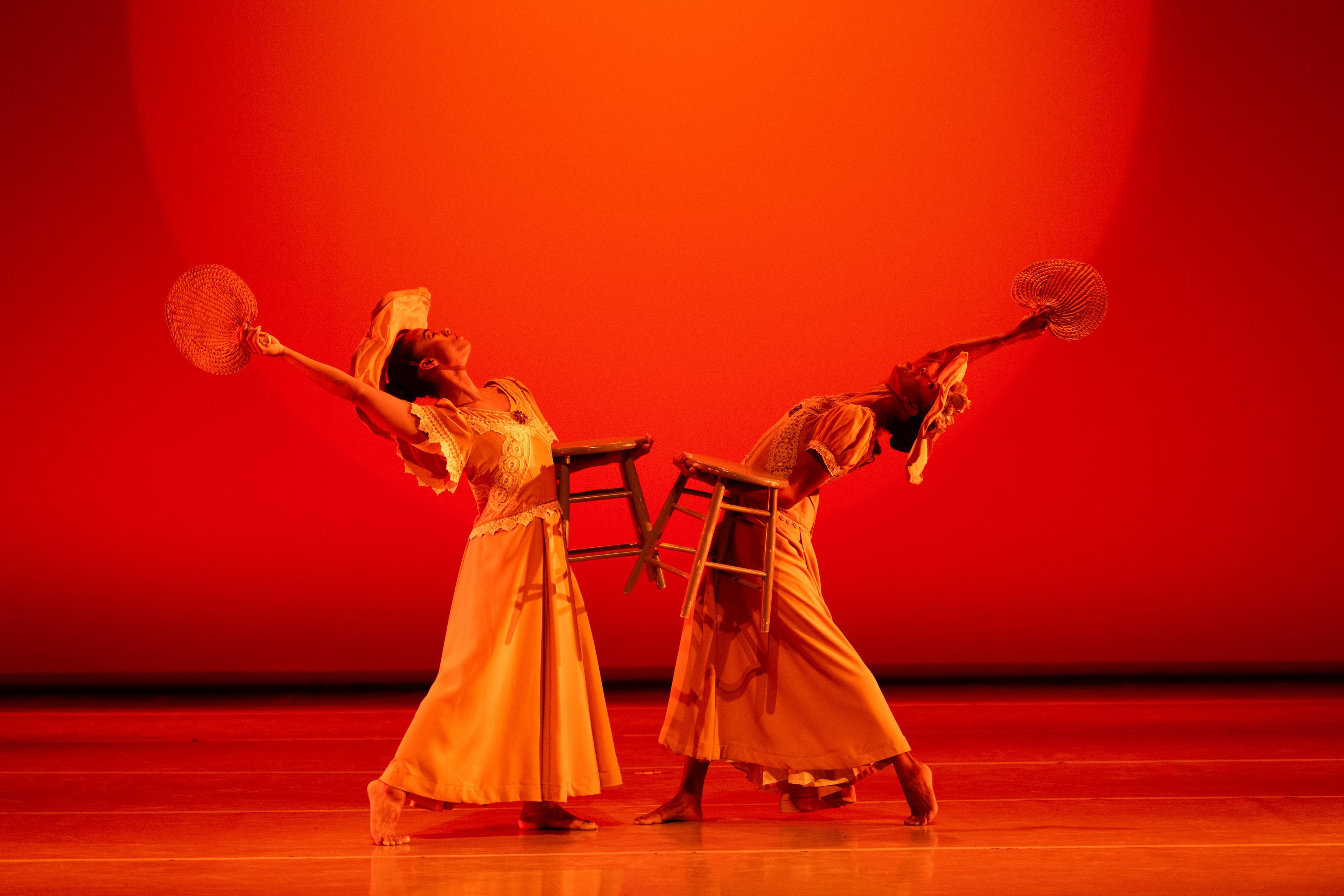 2 dancers in yellow arched away from each other holding a fan and a stool