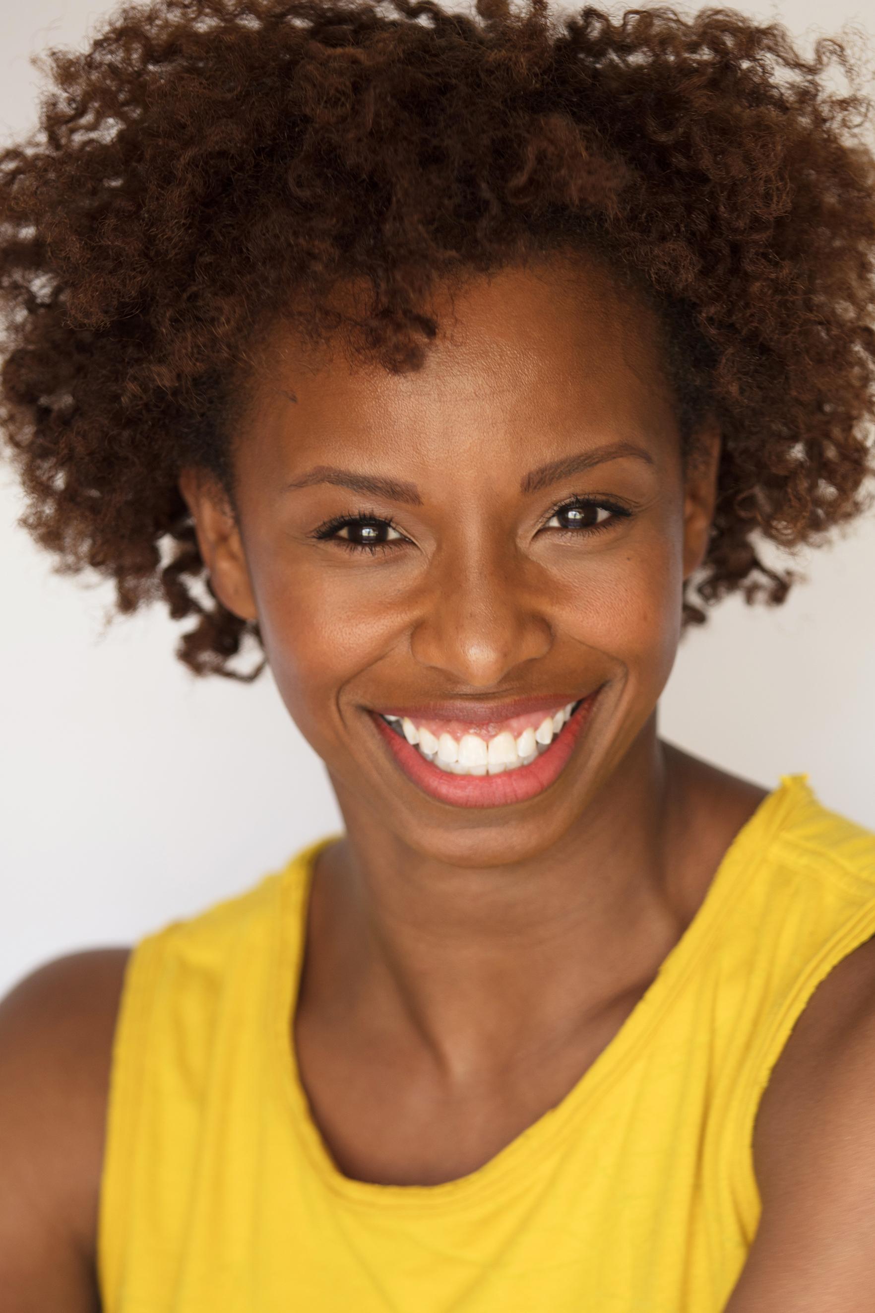 Portrait of Alia Kache wearing a yellow top. She is smiling brightly and looking directly at the camera. The background is plain and white.