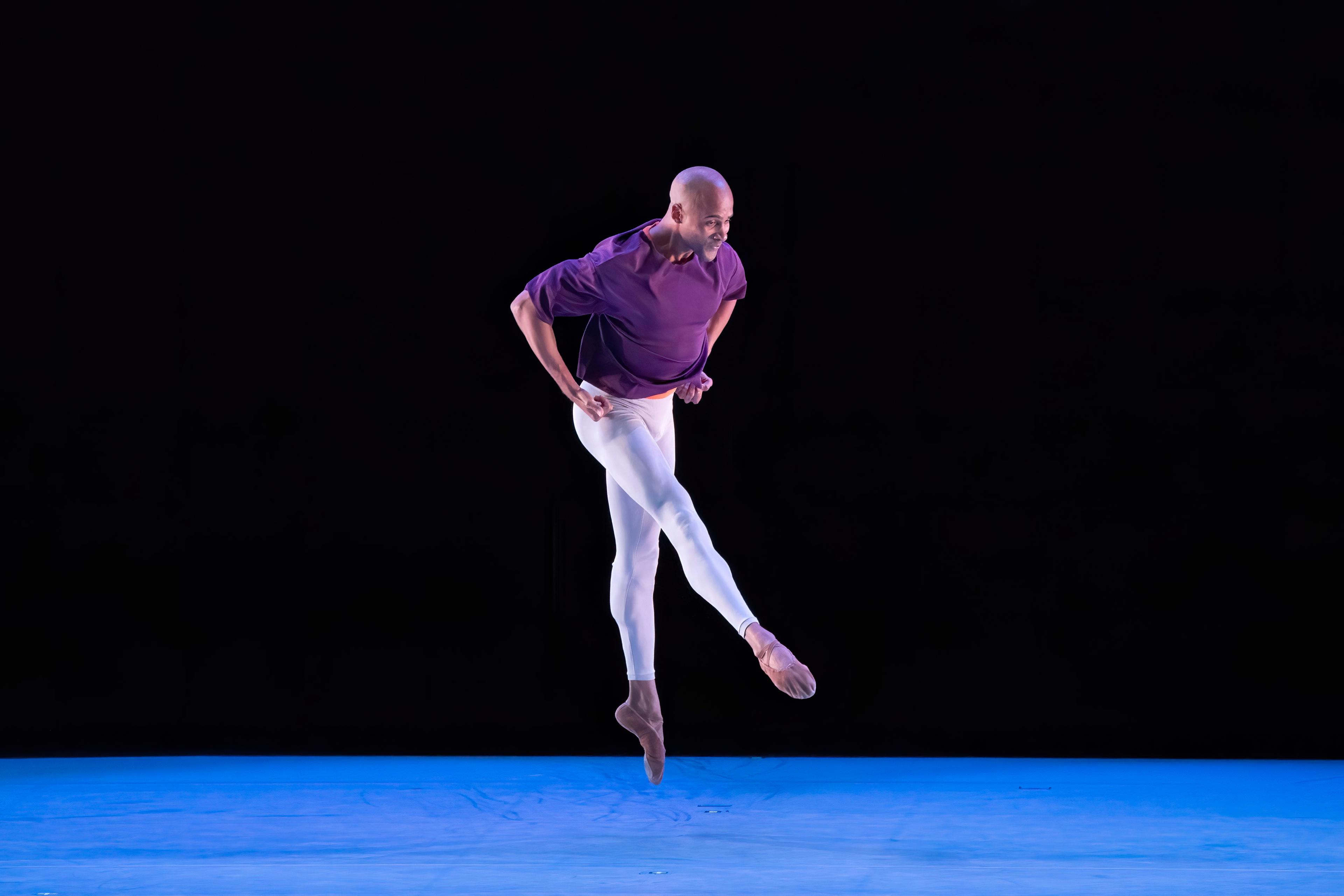 Dancer mid-jump. They are wearing a purple shirt and white pants. They are alone on stage.