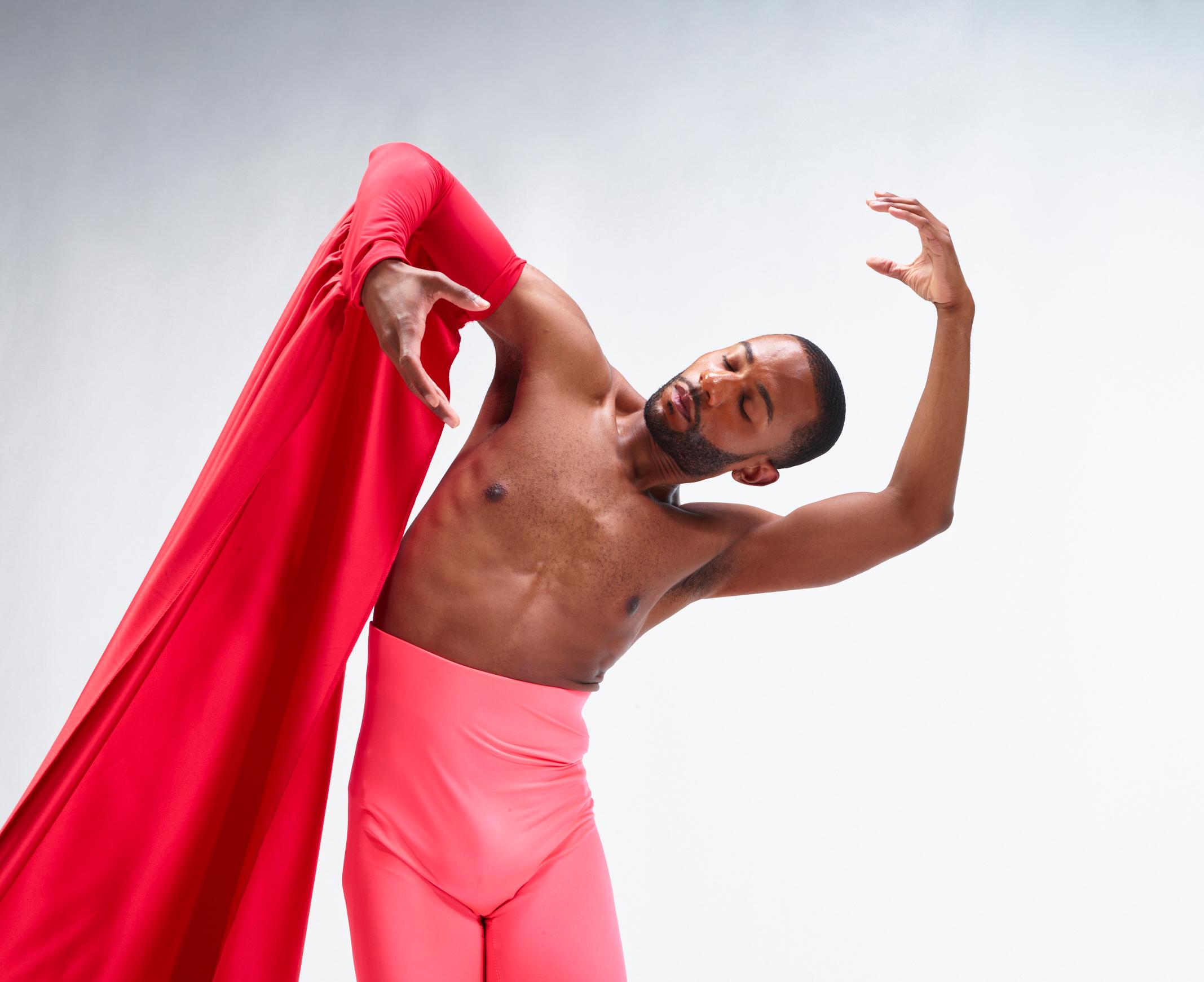 A shirtless male dancers leans to the right with red fabric draped over one arm.