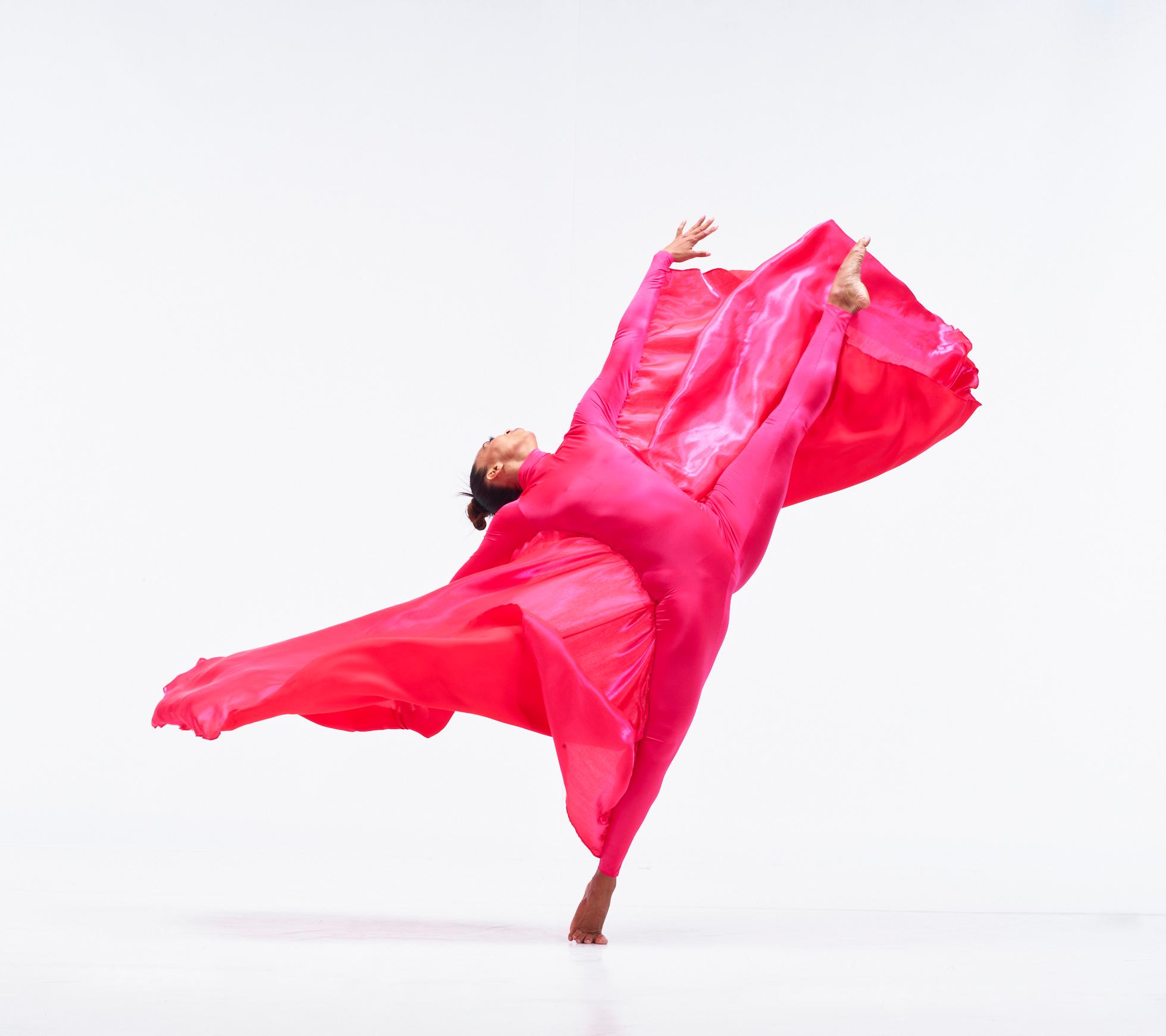 A dancer in a pink unitard and pink fabric kicks her leg up in the air