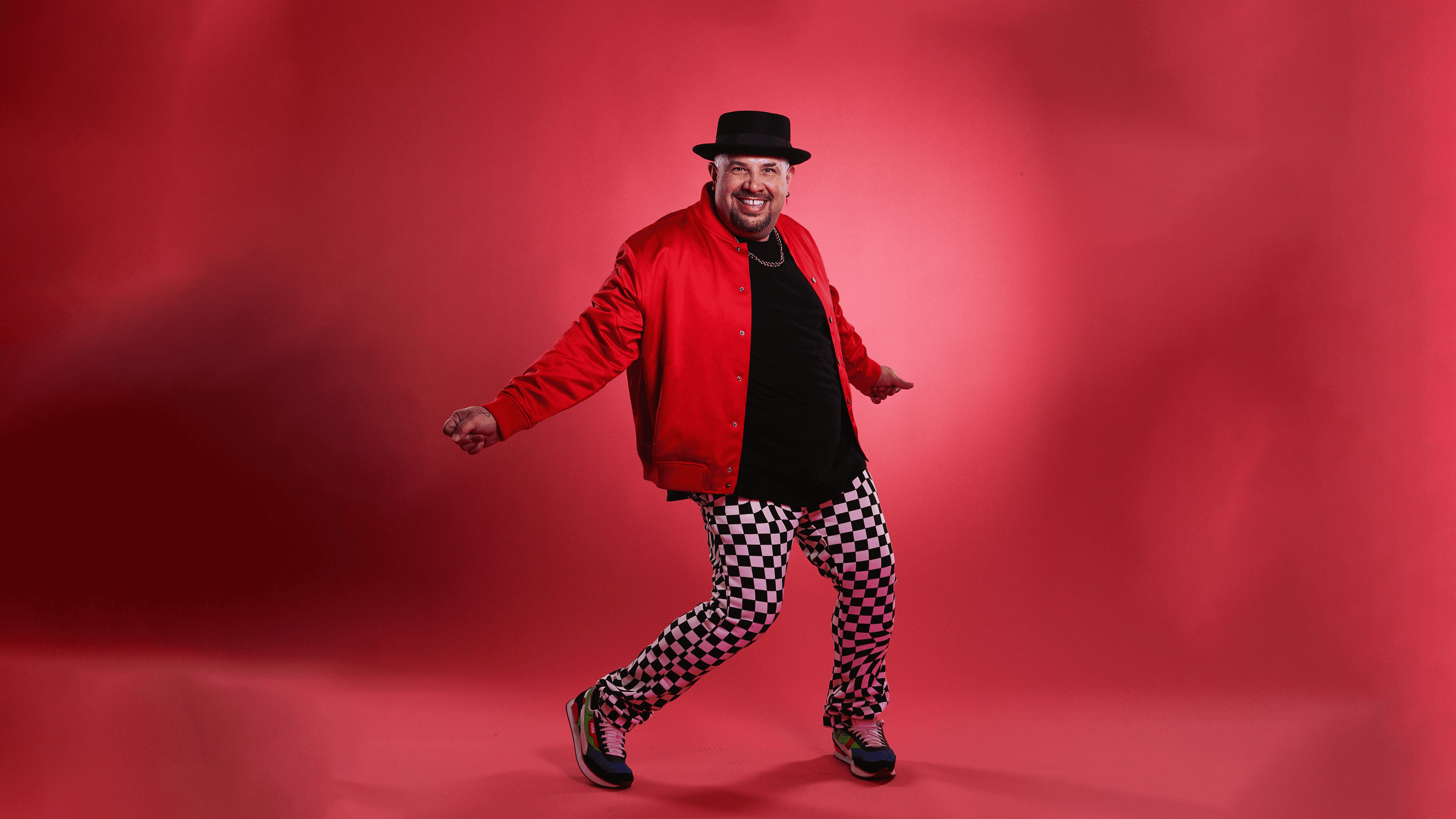 Lyrik Cruz dancing against a red backdrop wearing black and white checkered pants, a red jacket, and a black hat