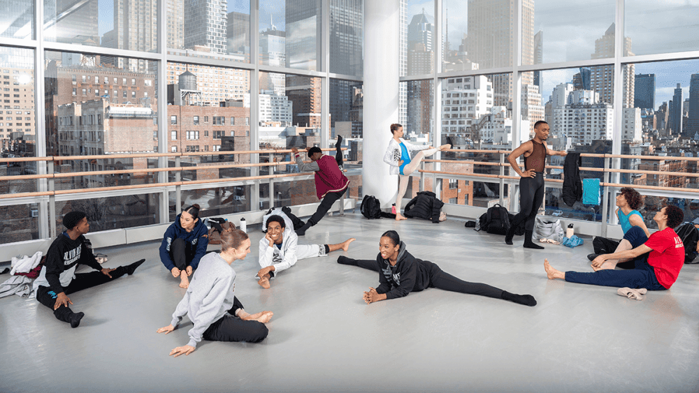 Students from The Ailey School Professional Division stretch and chat in a bright, spacious studio with large windows overlooking a cityscape. Some students are on the floor doing splits while others stand and stretch against the barre.