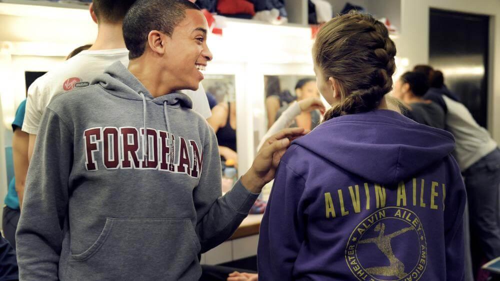 Ailey Fordham BFA Program students conversing in a dressing room, one wearing a Fordham hoodie and the other an Alvin Ailey hoodie. Photo by Eduardo Patino, NYC.