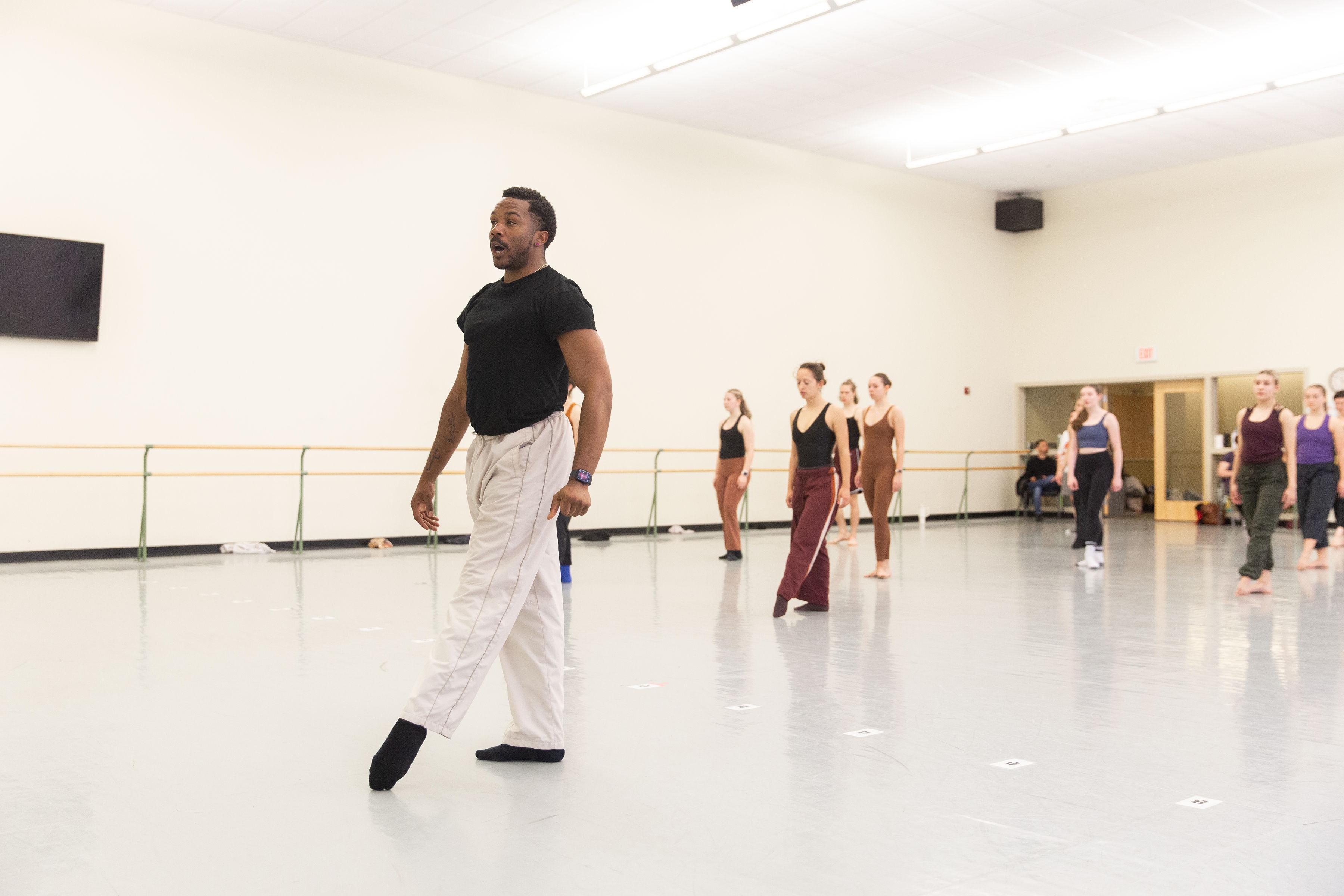 A male dance instructor in a black t-shirt and white pants demonstrates a move in a dance studio. He stands on one foot with the other leg extended, and students behind him follow along. The studio has light-colored walls, a ballet barre, and a TV mounted on the wall.