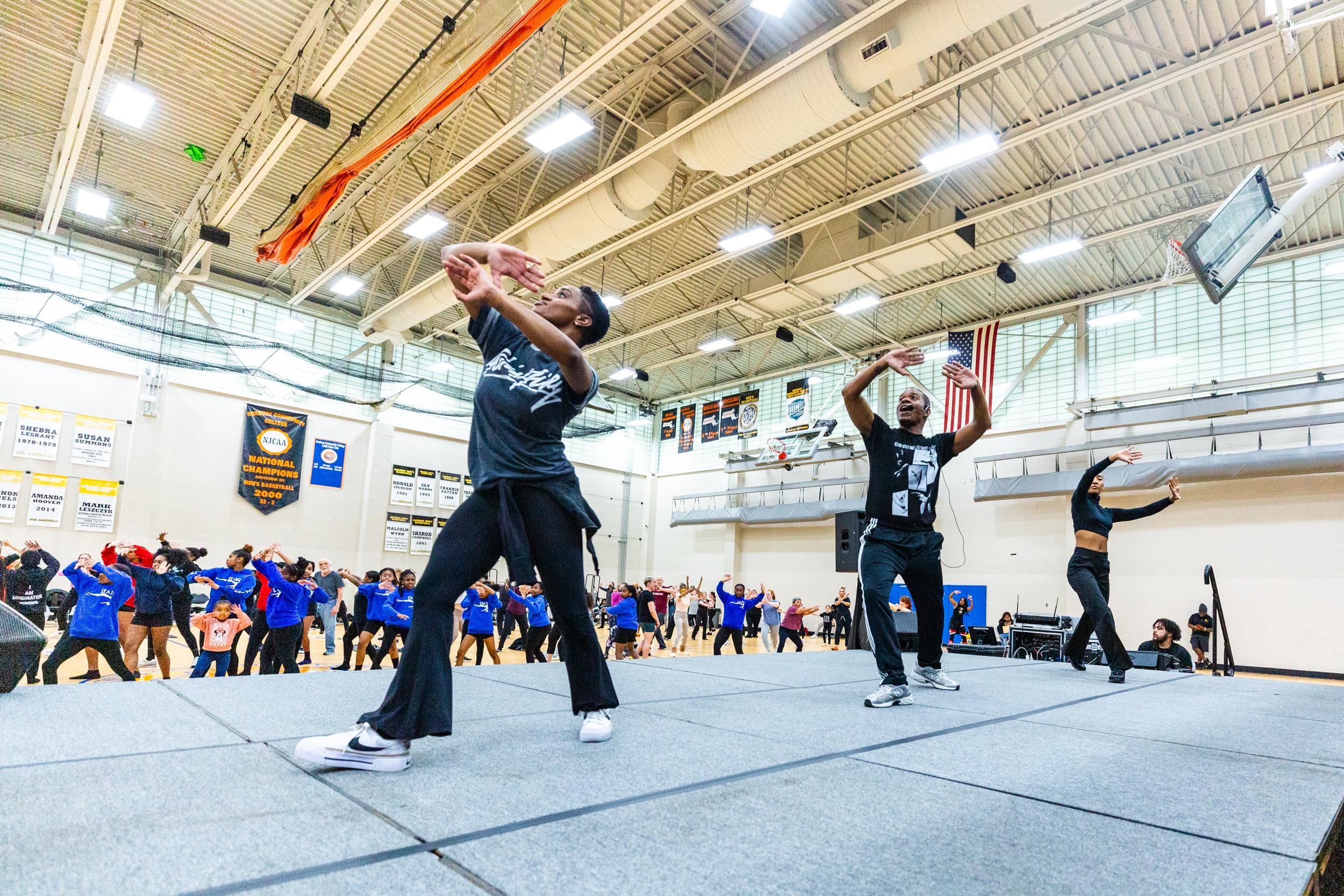 Three dance instructors on stage lead a large group of participants in a gymnasium. The instructors and participants are mid-motion, with arms raised. The gym is decorated with banners and flags, and the participants wear a variety of colors, mostly blue.