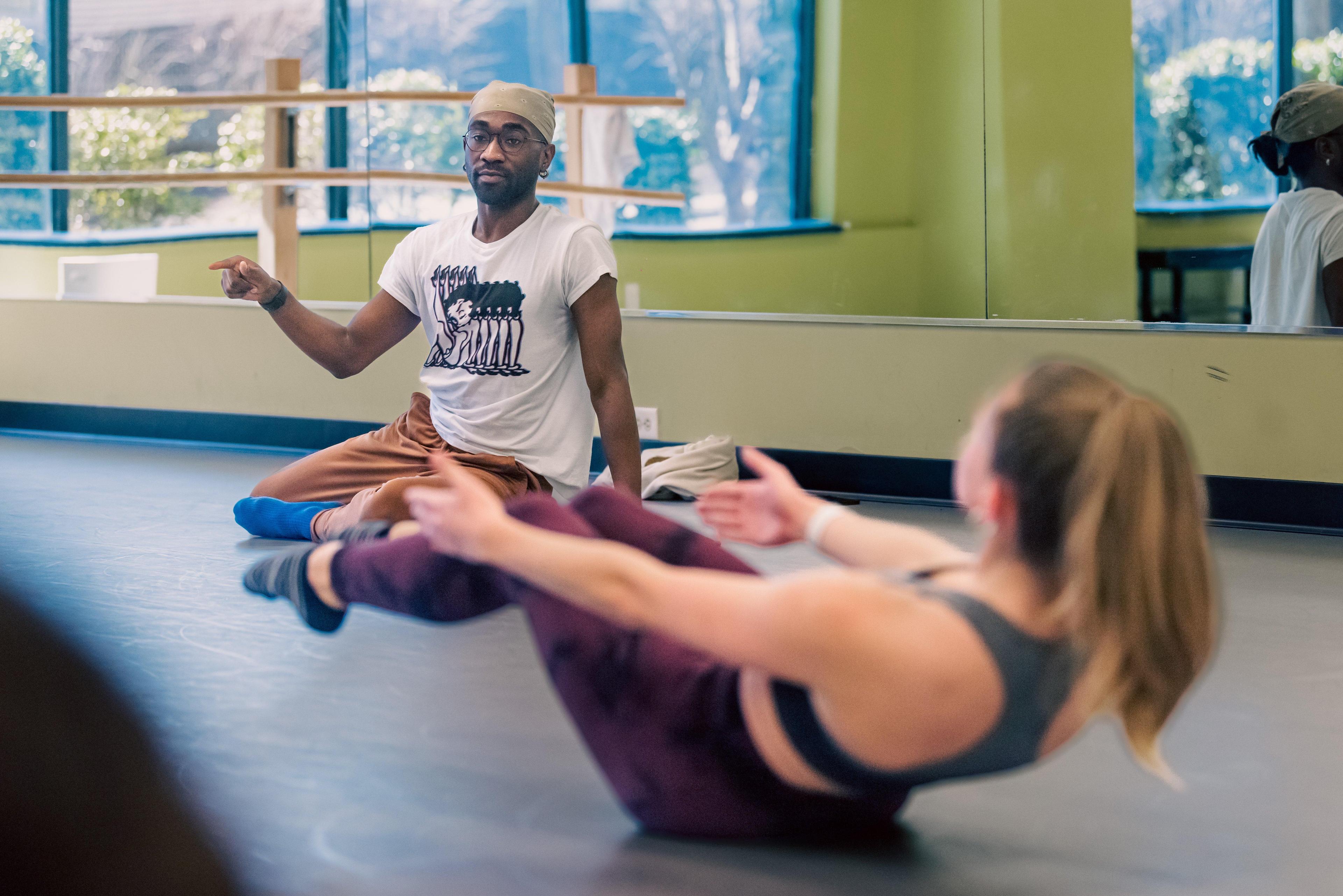 A male dance instructor wearing a headscarf, glasses, and a white t-shirt with a graphic, sits on the floor of a dance studio pointing to a student. The student, in focus, is performing a floor exercise in front of a mirrored wall. The studio has light green walls and large windows in the background.