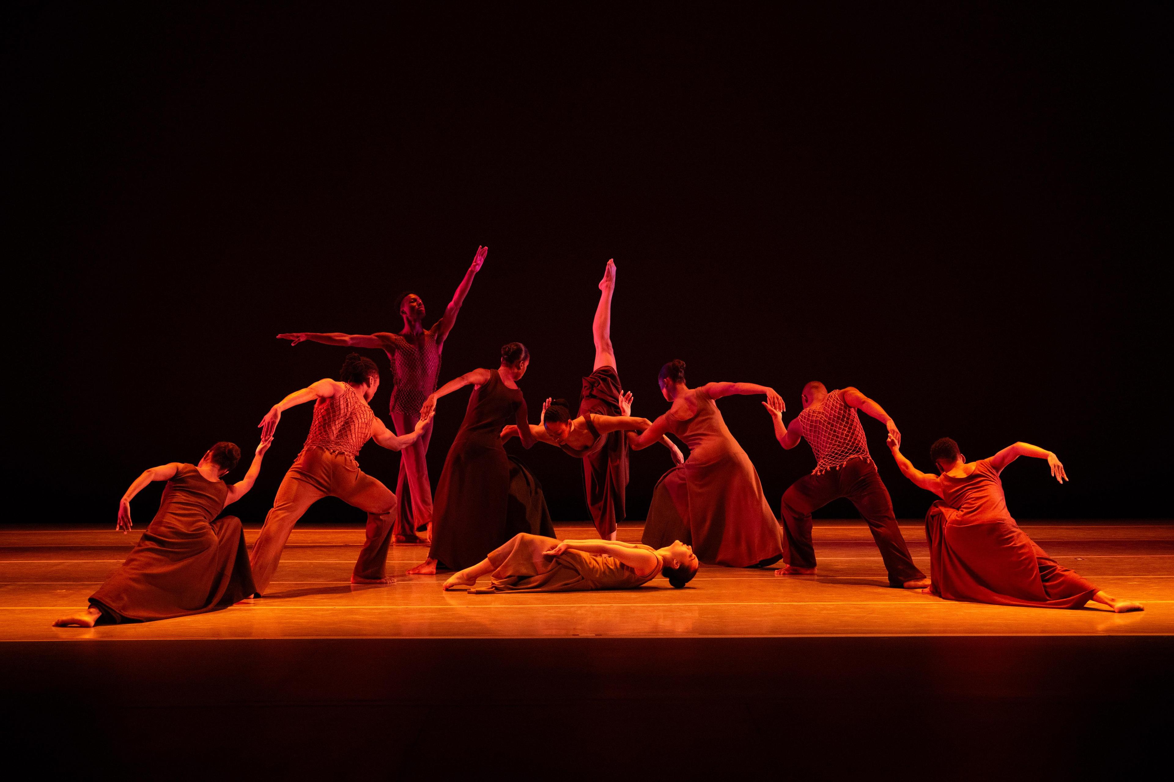 A group of dancers perform a dramatic routine on stage, illuminated by warm, reddish lighting. One dancer lies on the floor while others strike expressive poses, showcasing fluid and synchronized movements against a dark background.