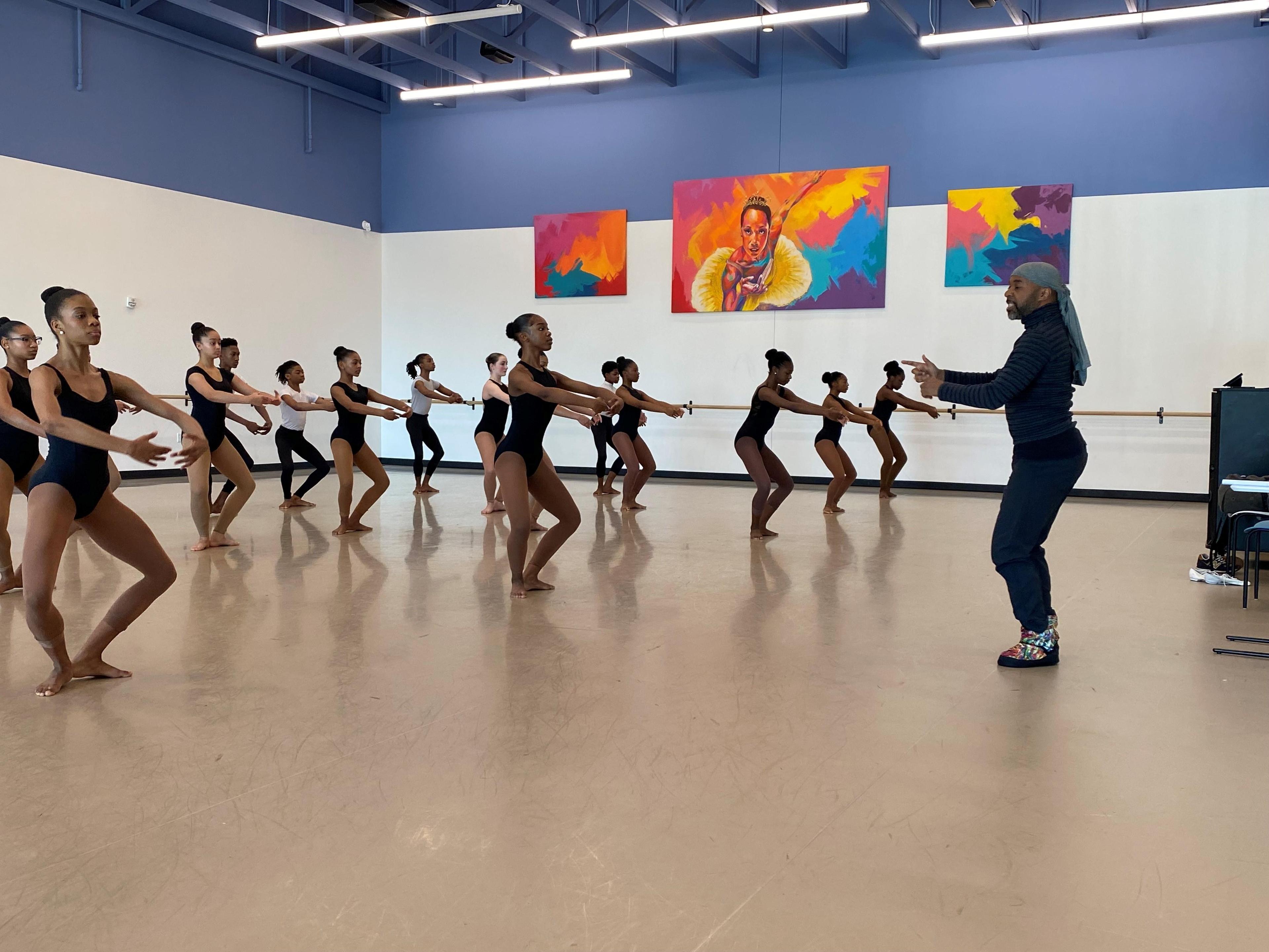 A group of ballet dancers in black leotards and tights, standing in a dance studio with light blue walls, taking instruction from a male dance instructor wearing a headscarf and dark clothing. There are colorful paintings on the back wall, and the floor is a light tan color.