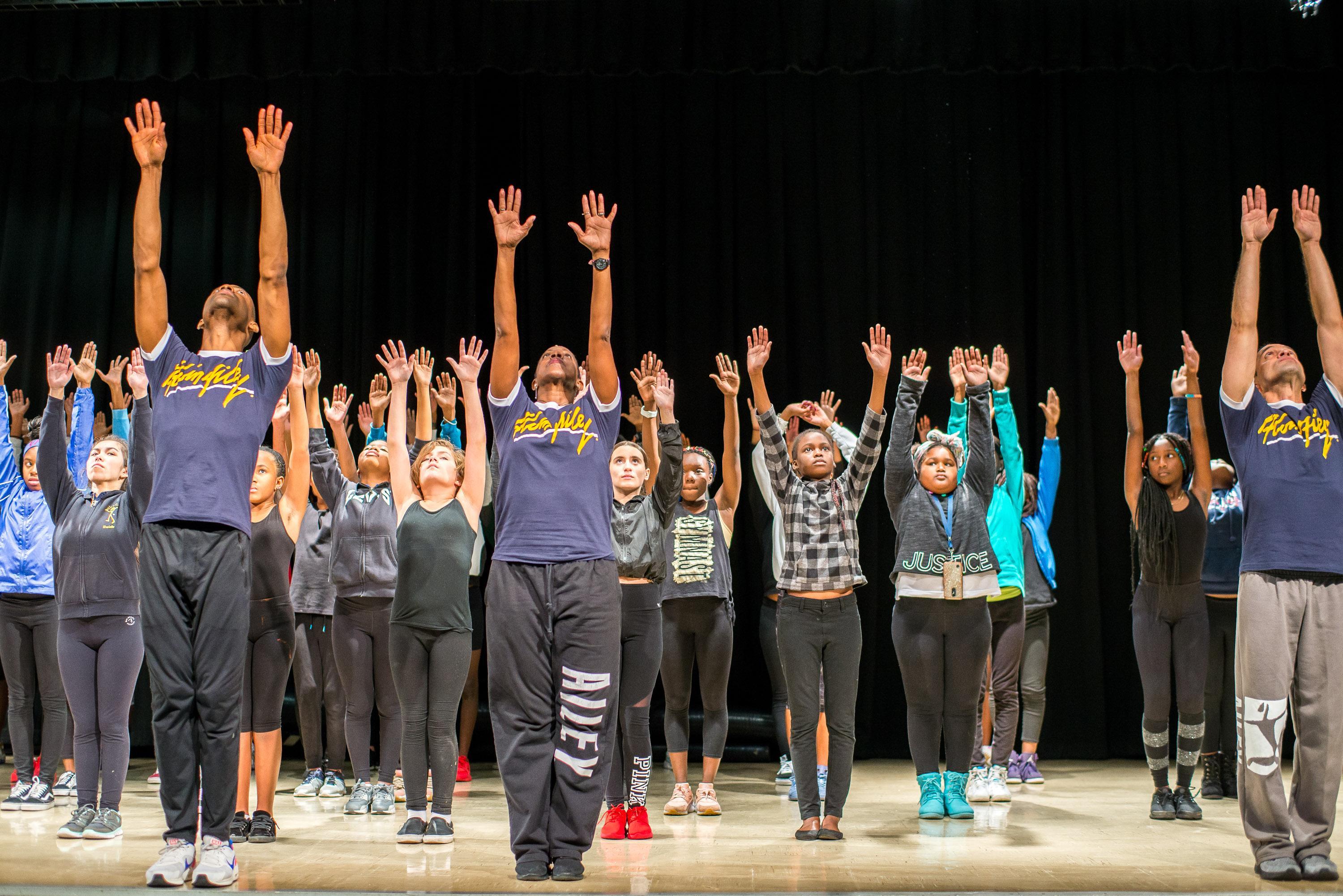  A group of dancers, including students and instructors in Alvin Ailey t-shirts, stand on stage with their arms raised high. They are performing a synchronized movement, all looking upward, showcasing unity and focus. The backdrop is a black curtain, emphasizing their poses.