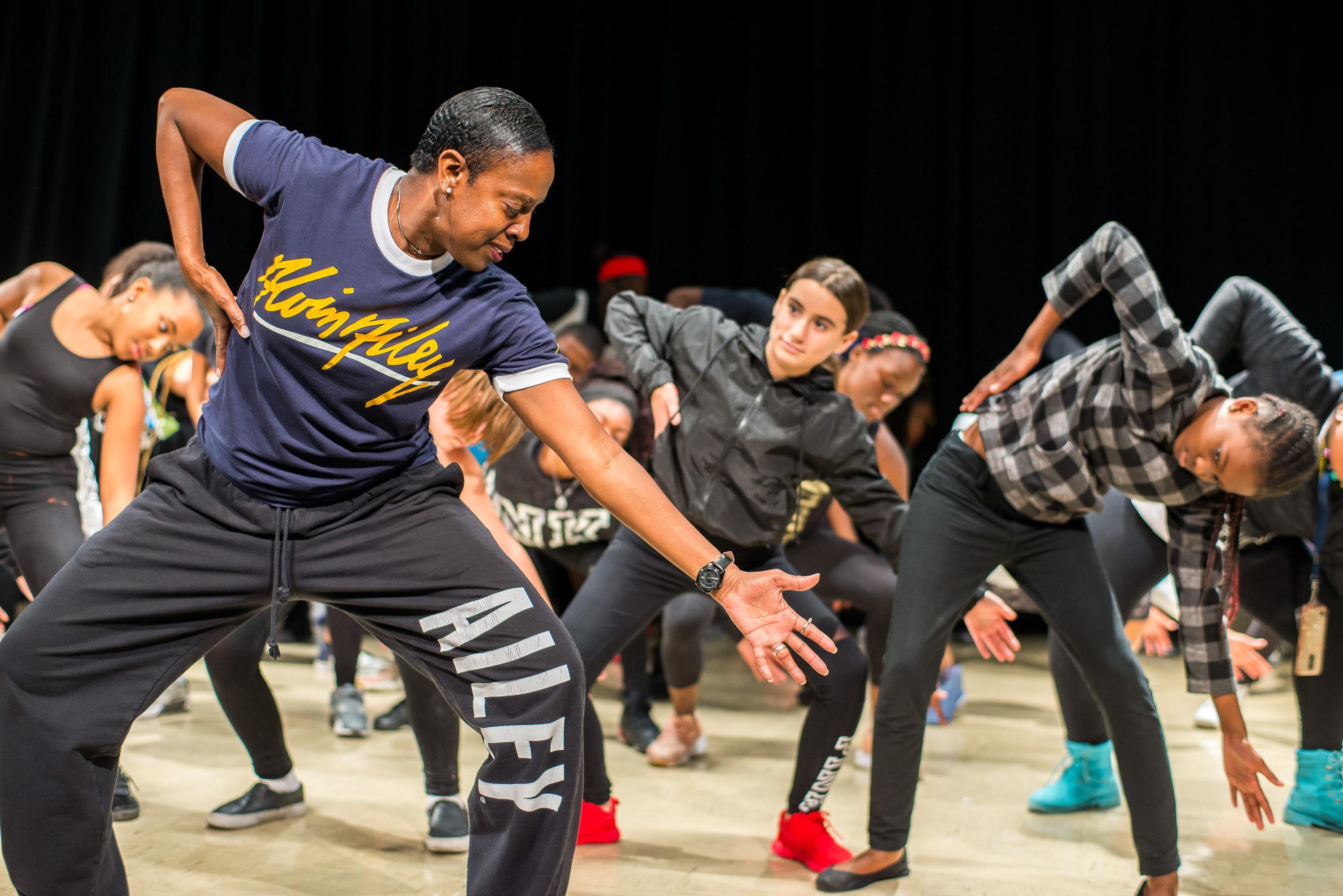 A dance instructor in a navy Alvin Ailey t-shirt and black sweatpants leads a group of students in a dance routine. The instructor strikes a low, dynamic pose, with the students behind mimicking the movement. The scene is energetic, with focused expressions and coordinated movements.