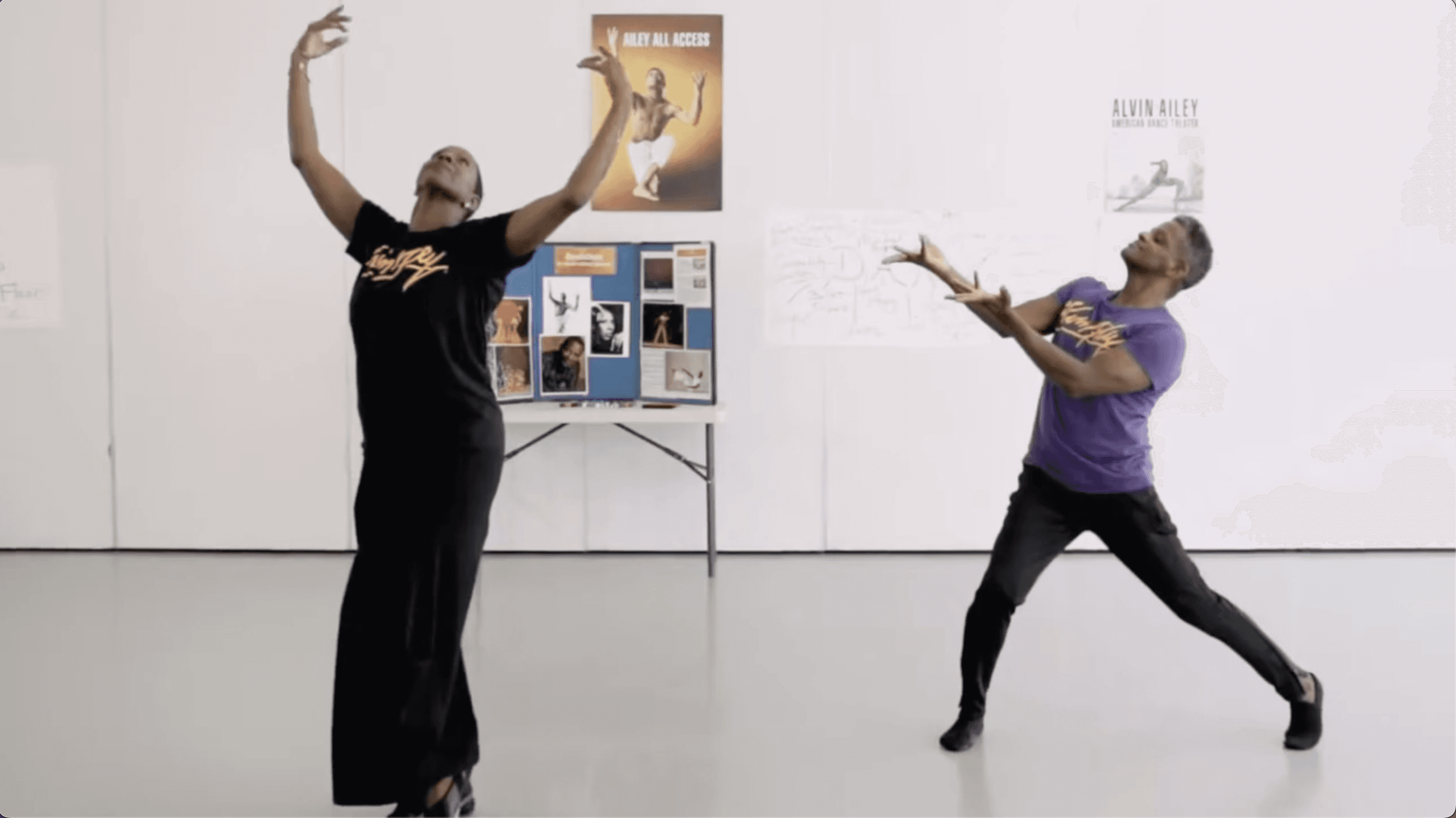 Two dancers perform in a studio, each striking a dramatic pose with arms extended. The dancer on the left wears a black t-shirt and long black skirt, while the dancer on the right wears a purple t-shirt and black pants. The wall behind them features posters and a display board with various images and text.