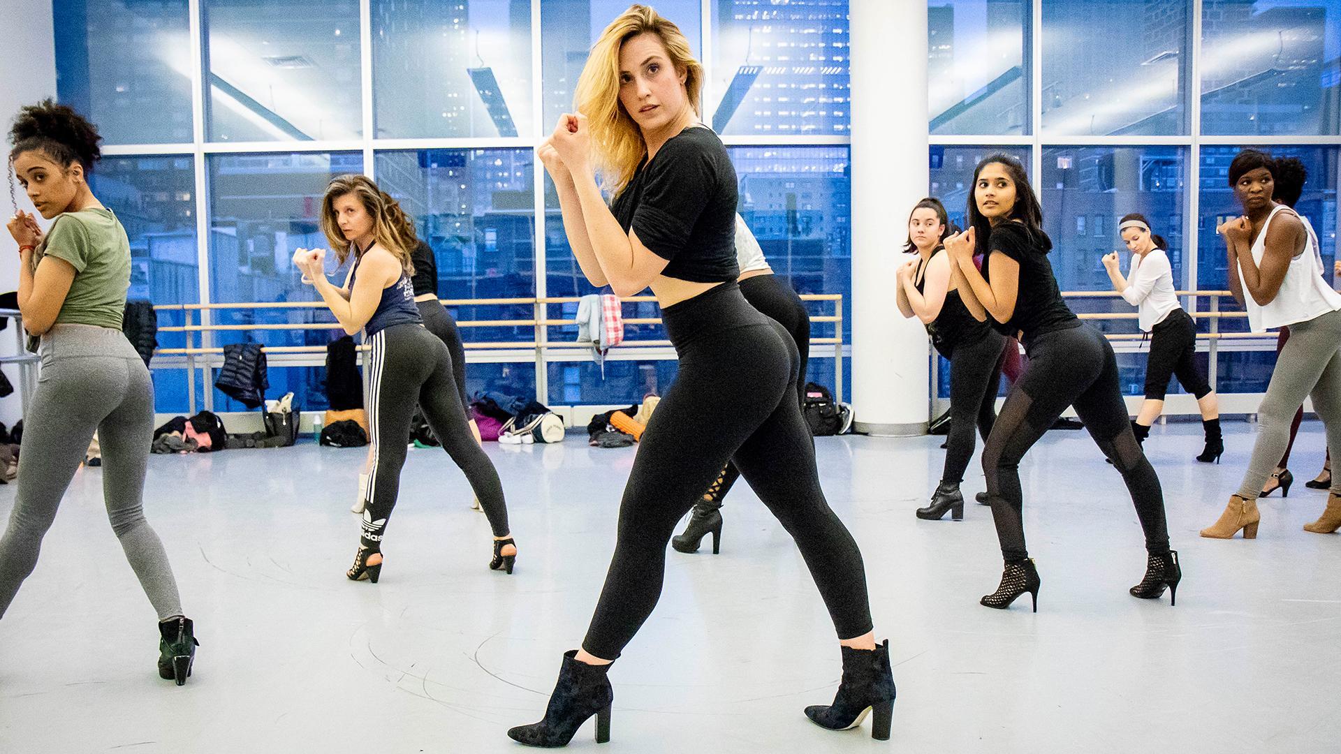 The dancers, dressed in stylish workout attire and high-heeled shoes, are captured in a powerful and confident pose, highlighting their strength and grace. The studio's large windows reveal a cityscape backdrop, adding an urban vibe to the energetic atmosphere. The instructor leads the class with precision, encouraging the dancers to embody the fierce and empowered style characteristic of Heels dance. 