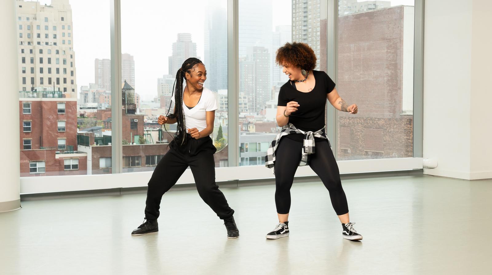 Both dancers display a sense of joy and energy as they engage in the dynamic and rhythmic style of House dance. The studio's large windows provide a clear view of the cityscape, adding to the urban feel of the dance style. The dancers' outfits, consisting of comfortable, casual clothing, highlight the street dance origins of House. 