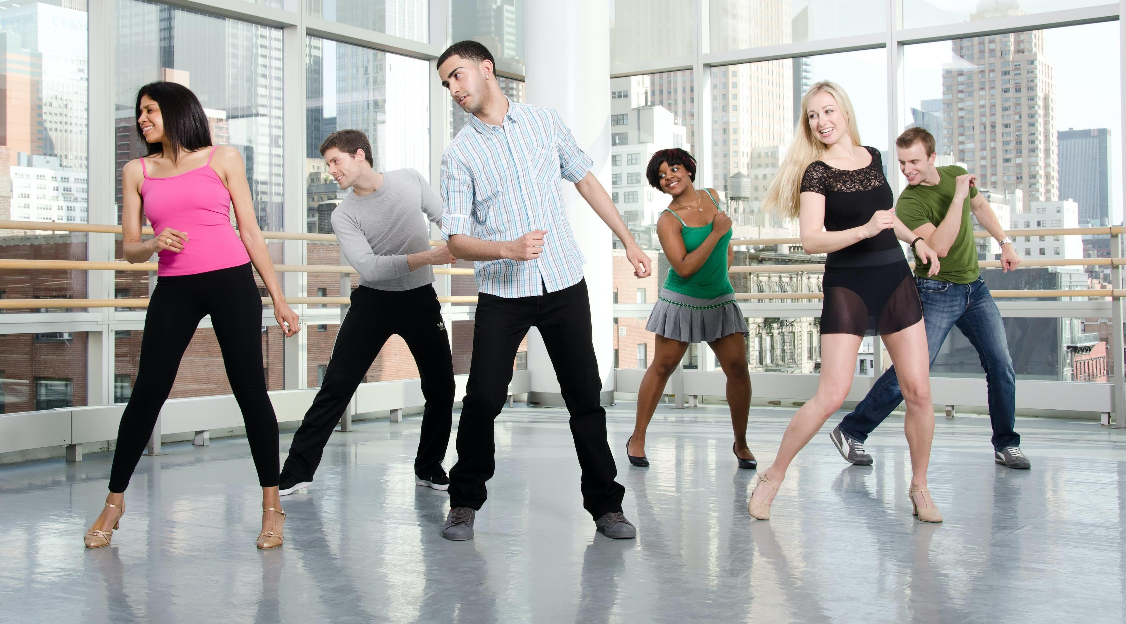 The image captures a vibrant Salsa class at the Ailey Extension. Participants, dressed in a mix of casual and dance attire, are engaged in learning and practicing salsa dance moves. The diverse group of men and women are shown mid-movement, displaying enthusiasm and enjoyment. 