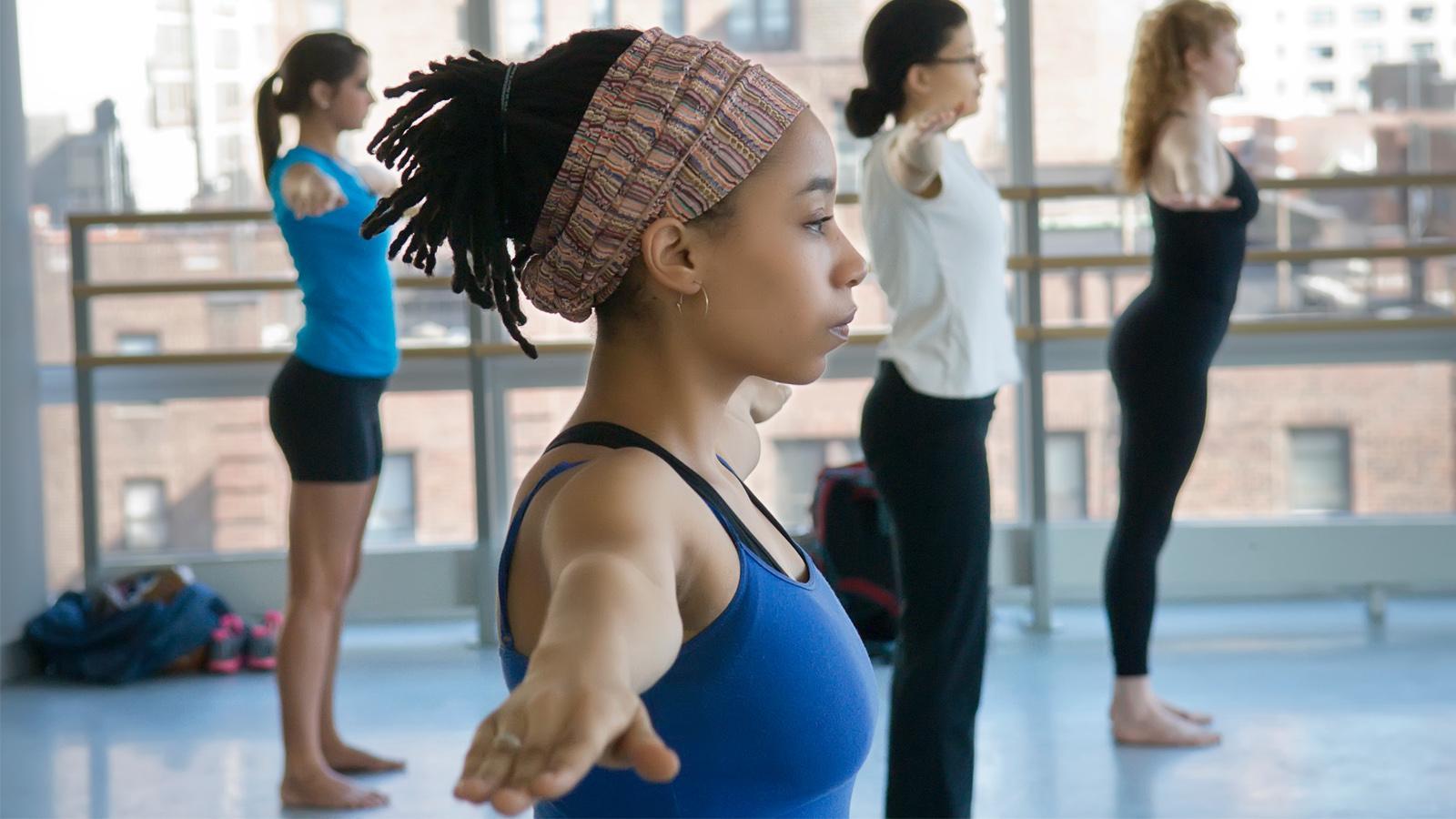 The dancers, dressed in comfortable workout attire, are deeply engaged in their movements, embodying strength and grace. The woman in the foreground, with a headband and neatly styled hair, stands out with her determined expression and  poised stance. The class environment, featuring large windows and a bright, airy studio, adds to the sense of energy and concentration. 