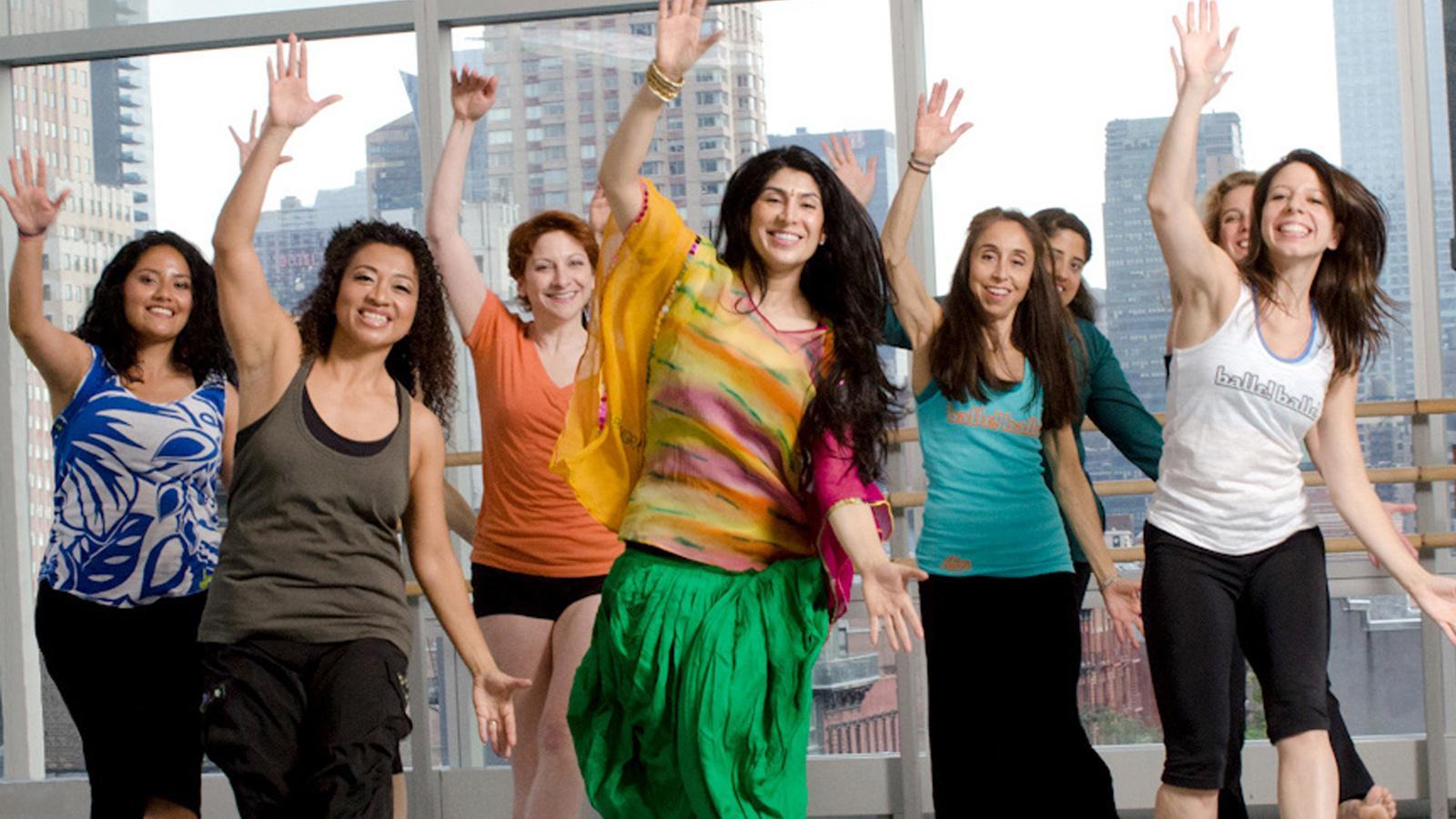 The participants, a diverse group of enthusiastic dancers, are engaged in a Masala Bhangra class, characterized by its high-energy movements and joyful expressions. The instructor, dressed in colorful attire, leads the class with dynamic gestures, encouraging everyone to follow along with enthusiasm. The spacious studio, filled with natural light from large windows, provides an inspiring backdrop that enhances the overall atmosphere of fun and community. 