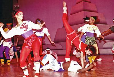 Dancers dressed in red silk pants and crop white sweaters are posed on a dance floor.