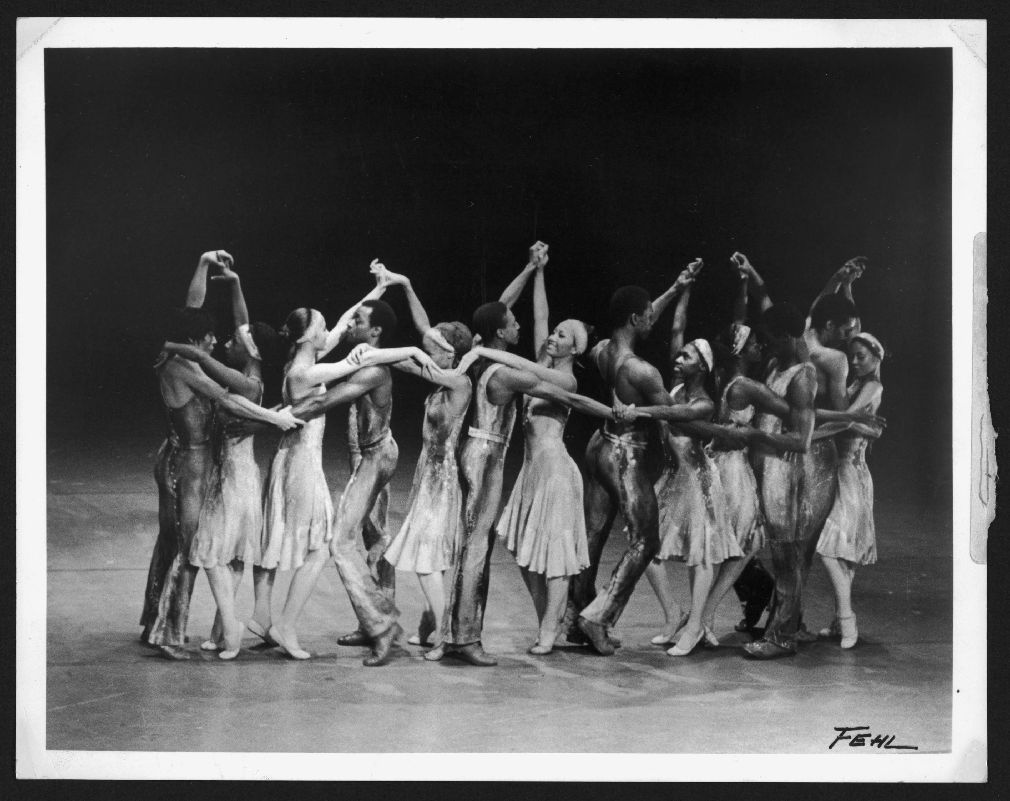 Black and white photo of Alvin Ailey dancers in a performance. The dancers are arranged in a circle, holding each other's arms, wearing flowing costumes. The background is dark, highlighting the dancers. Photographer's signature, Fehl, is visible in the bottom right corner.