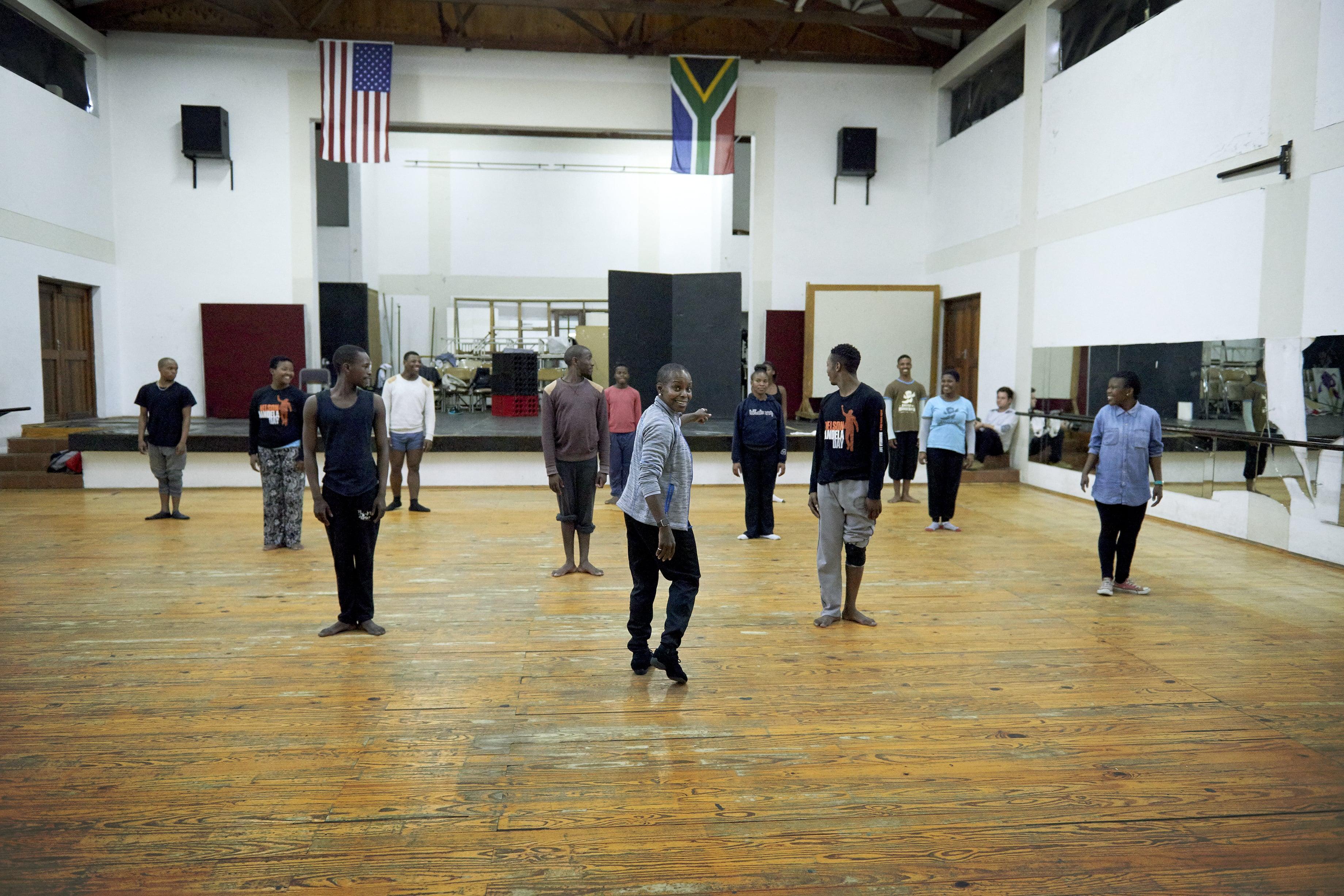 Group of dancers in a studio with wooden floors and white walls. The dancers are spaced apart, following the instructions of the lead dancer in the center, who is gesturing. American and South African flags hang on the back wall. Mirrors are on the right wall. 